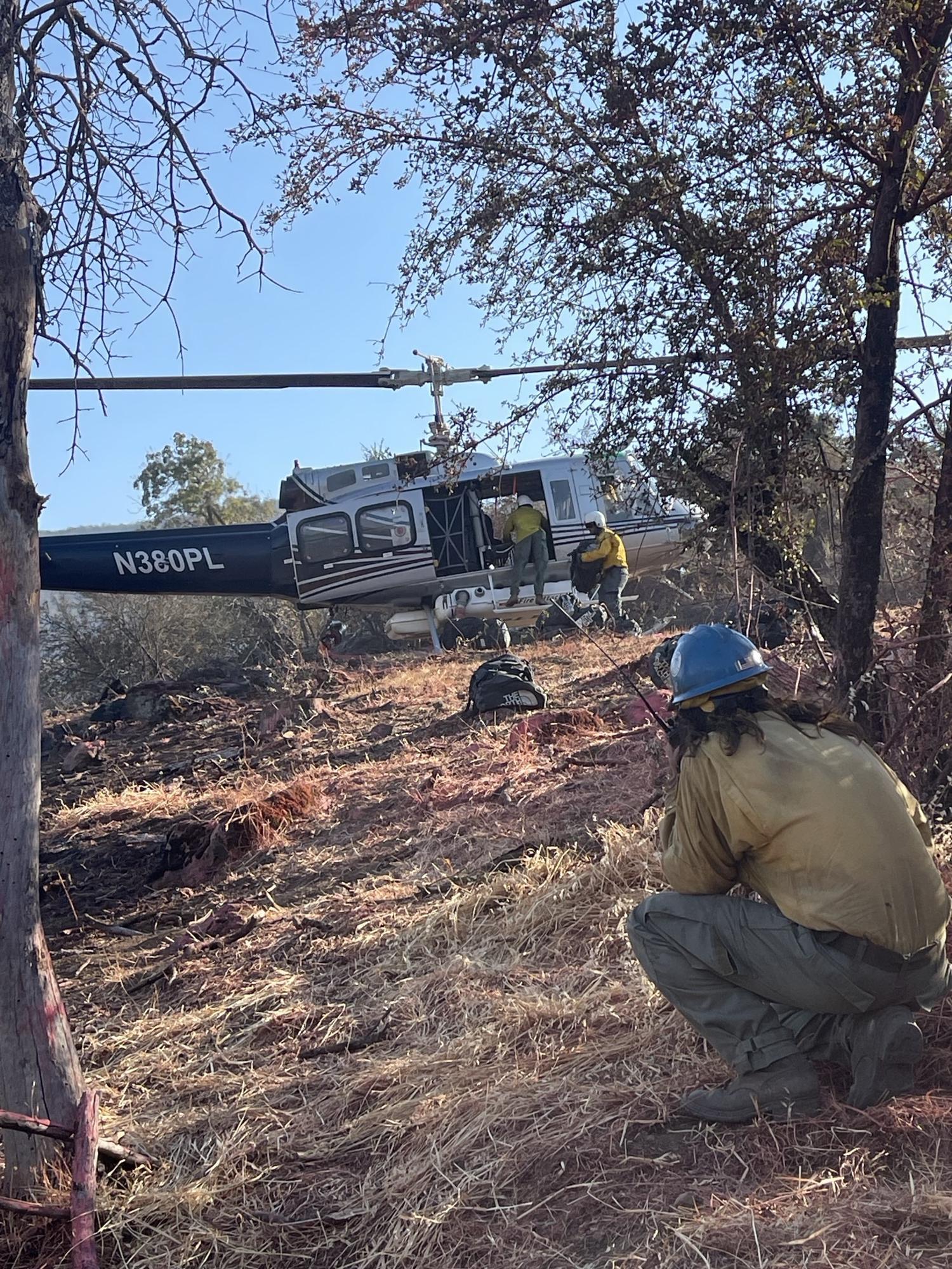 A firefighter kneels in the foreground talking on a radio. He is looking at a helicopter as two more firefighters unload supplies.