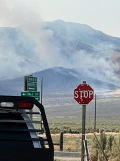 Smoke rises over a mountain range, with a road, stopsign and portion of a truck in the foreground