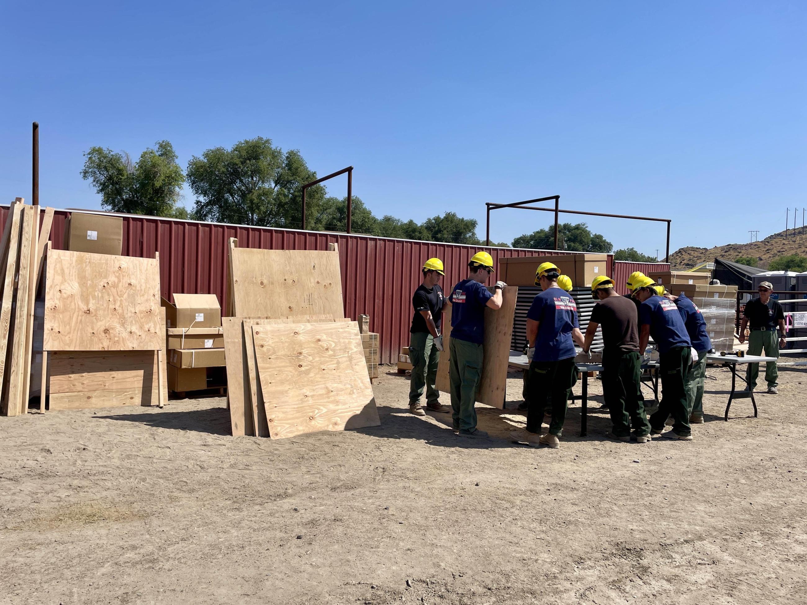 A crew of people work around a table outside, with finished wooden "easel-style" wooden boards to their left