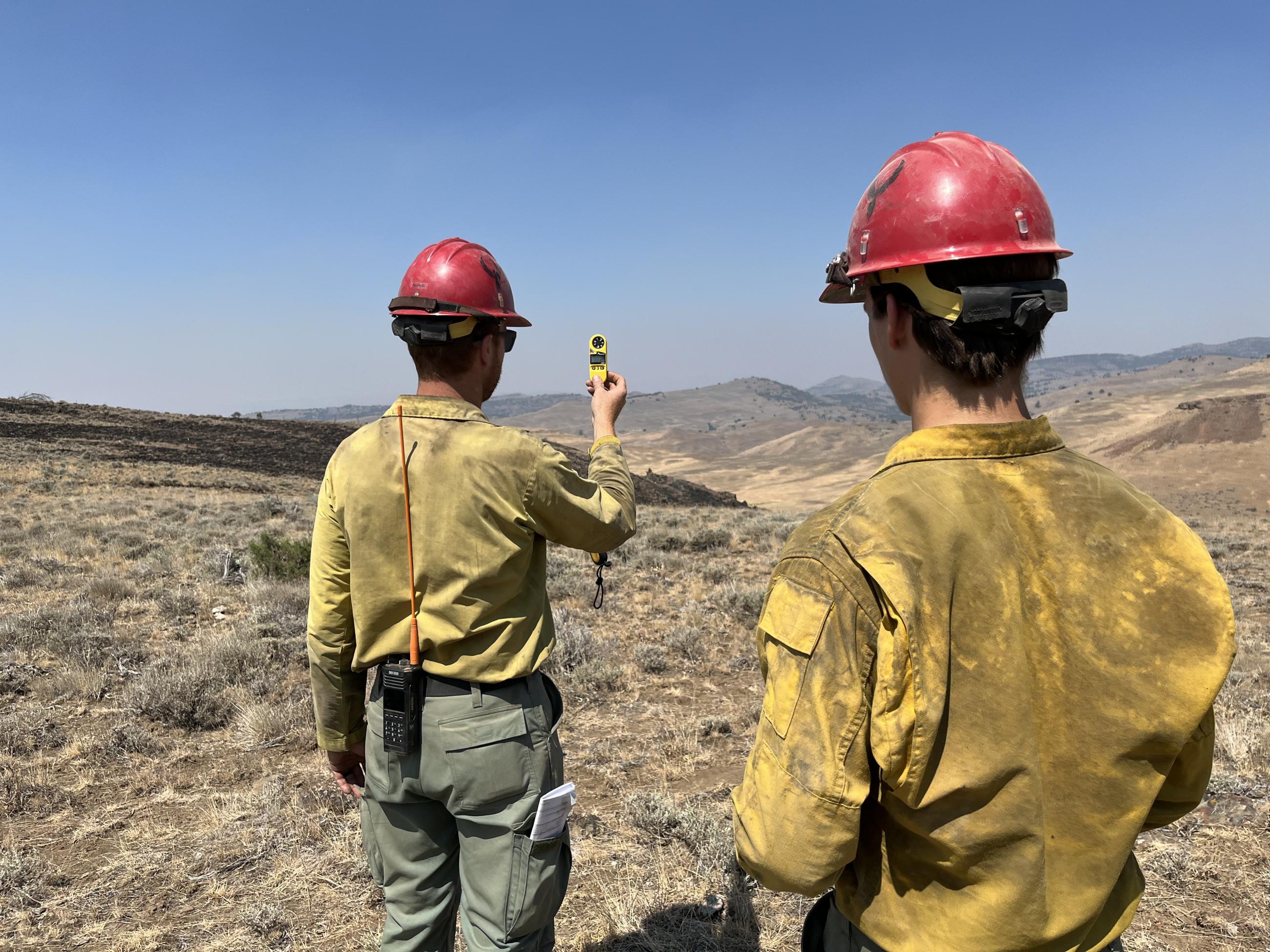 Two firefighters holding a small instrument up, shrubland in the background