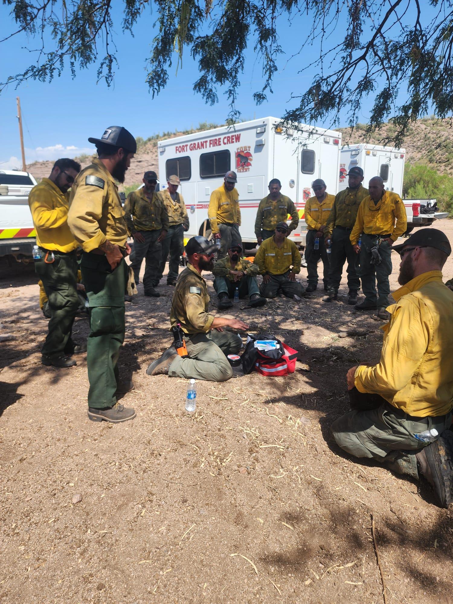A team of medical professionals in firefighting yellows gather in a circle around an instructor who is speaking while on his knees in the middle of the group. 