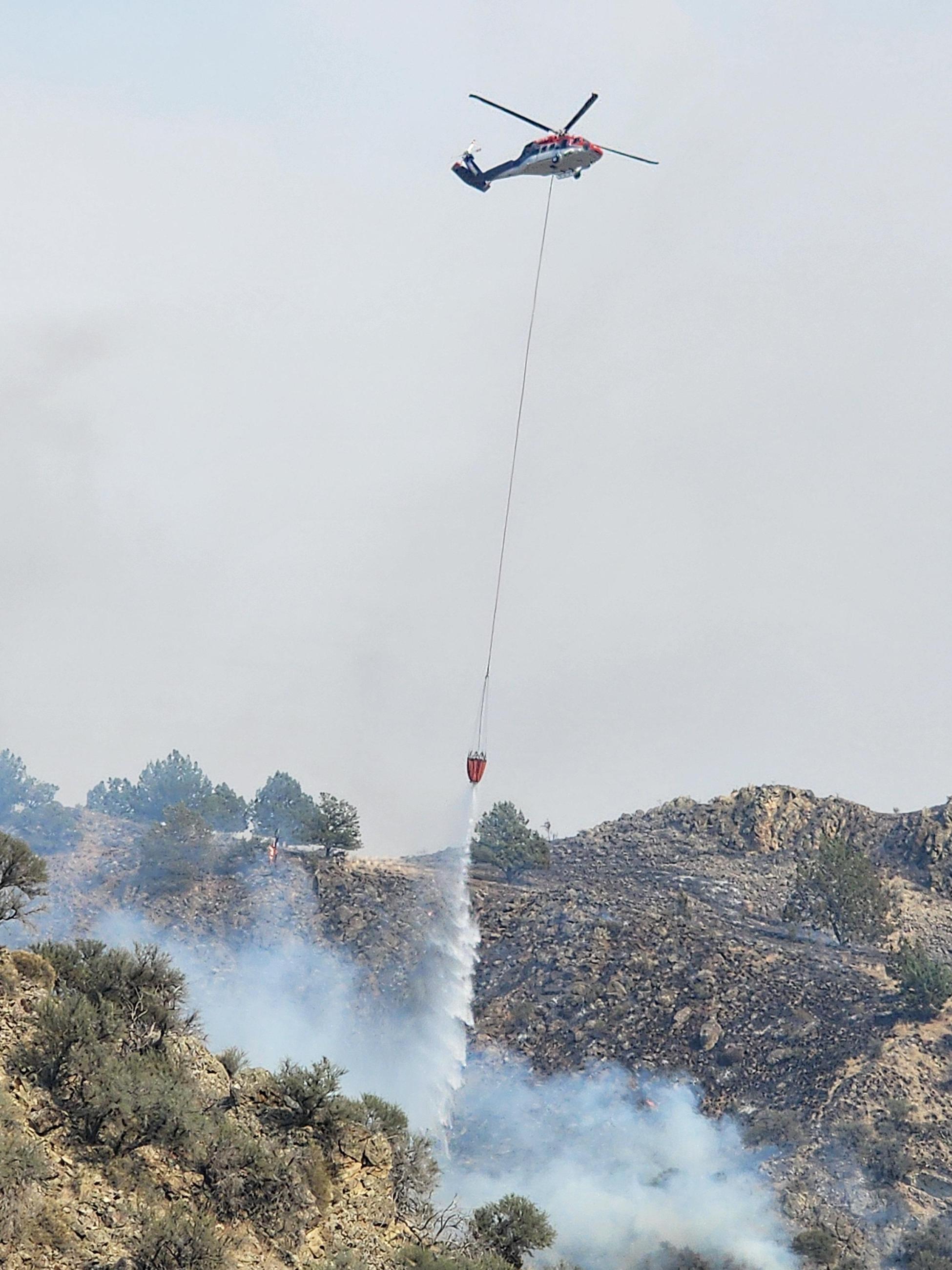 Helicopter flying in a smoky sky with a line attached to a bucket dropping water in a smoke filled canyon