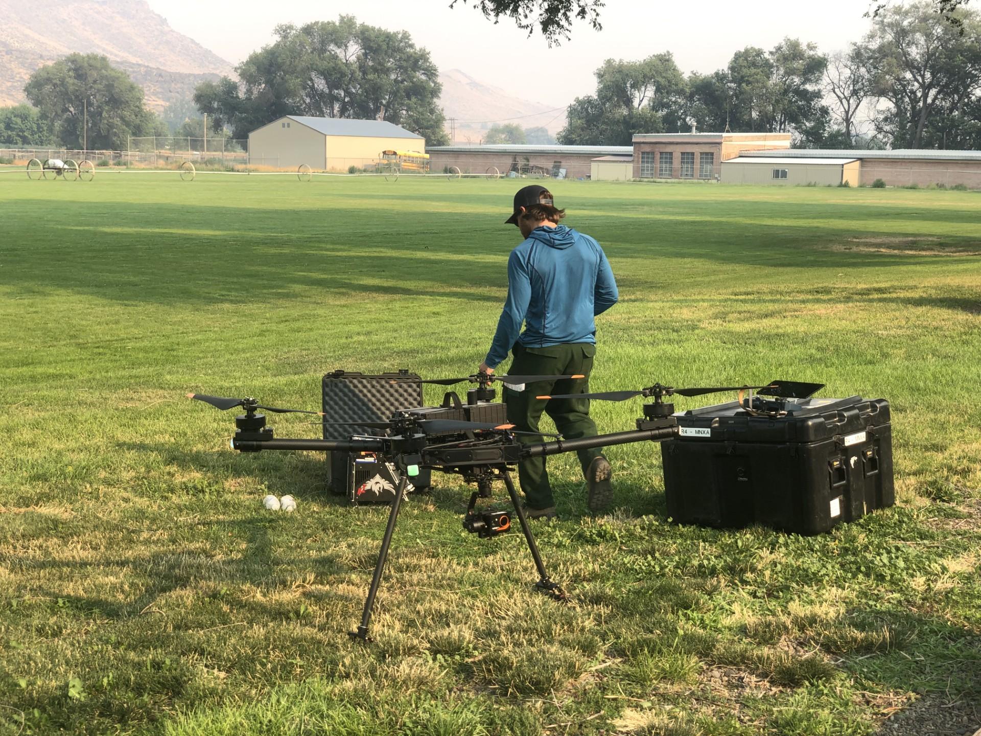 Drone and operator in a green field with buildings, trees, and hills in the distance