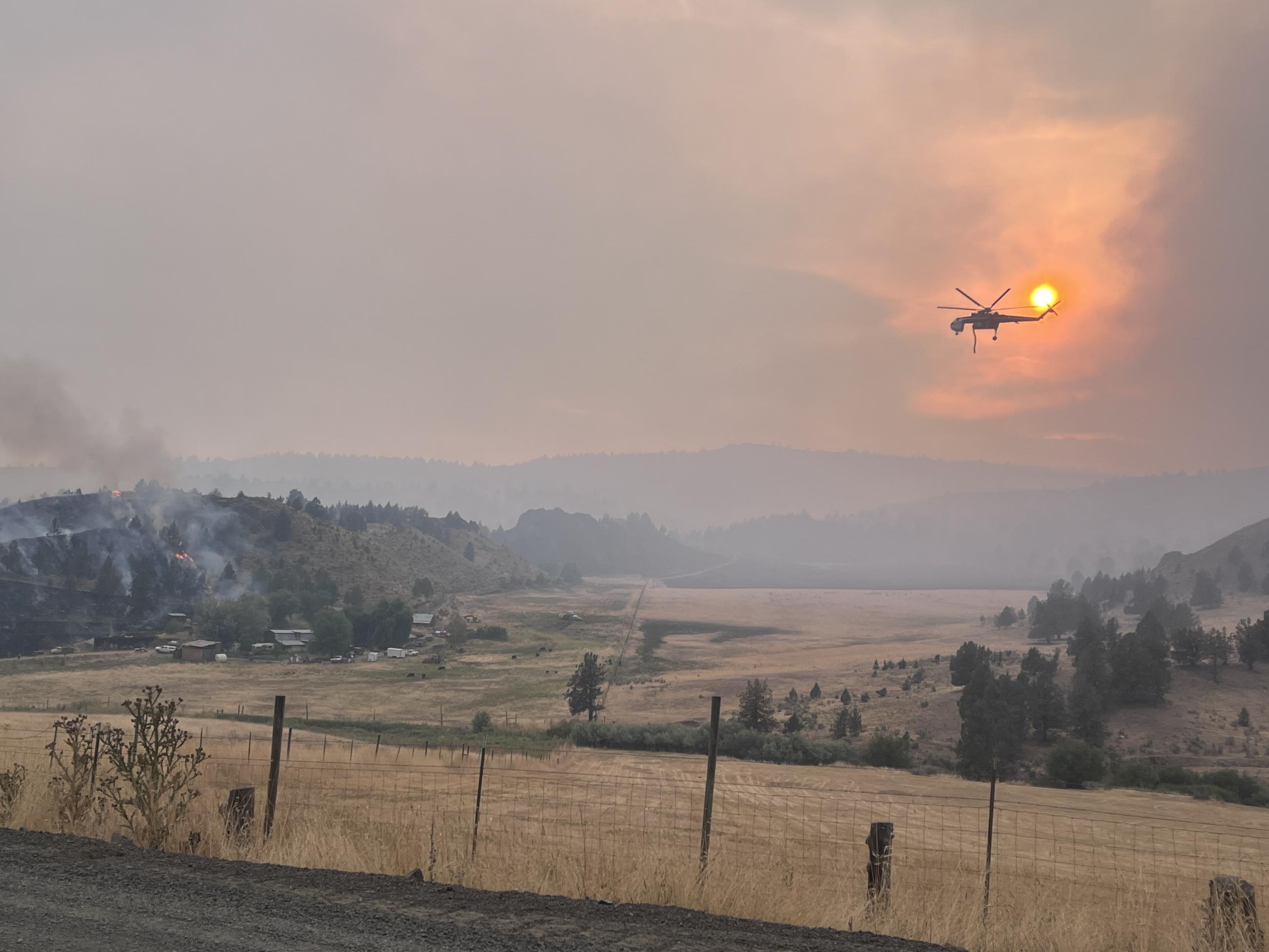 Image of a helicopter flying through a smoky sunset with a field and bluffs below.