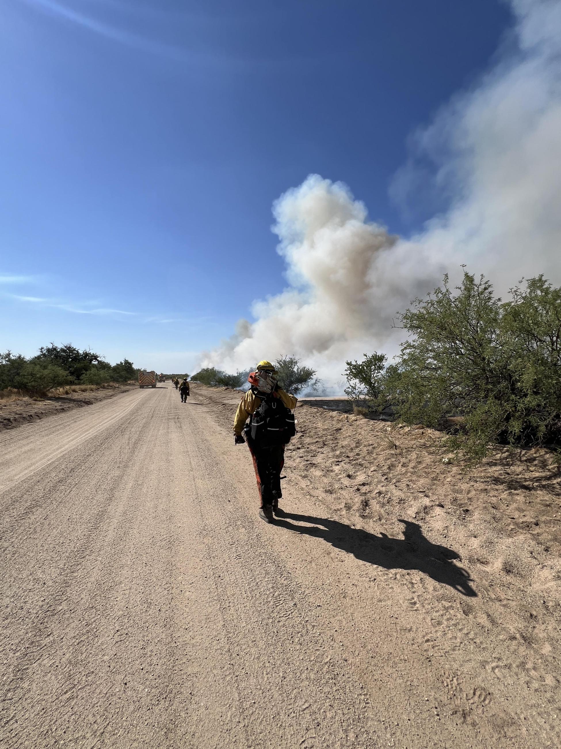Firefighter carrying chainsaw walks down dirt road with smoke rising in background against blue sky