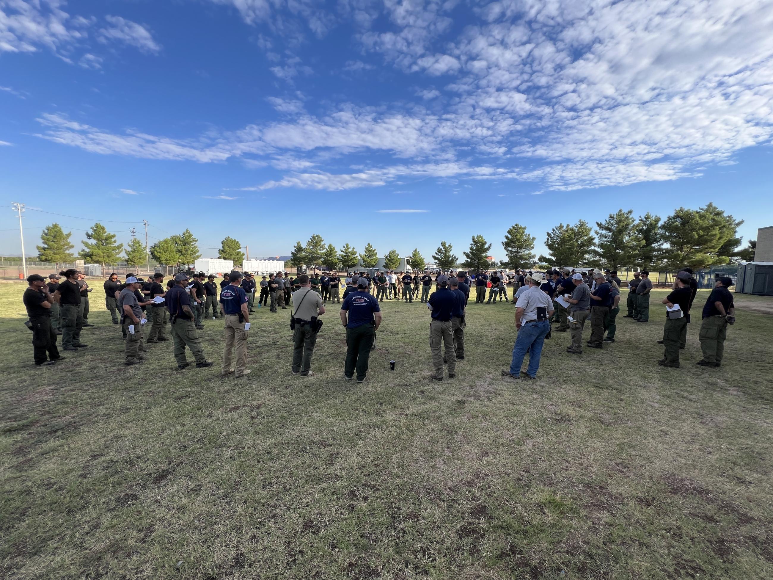A couple dozen people stand in a circle on grassy field to get an update on operations