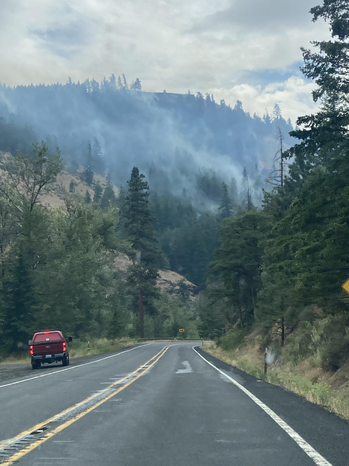 A highway in a timbered canyon with smoke rising from trees.