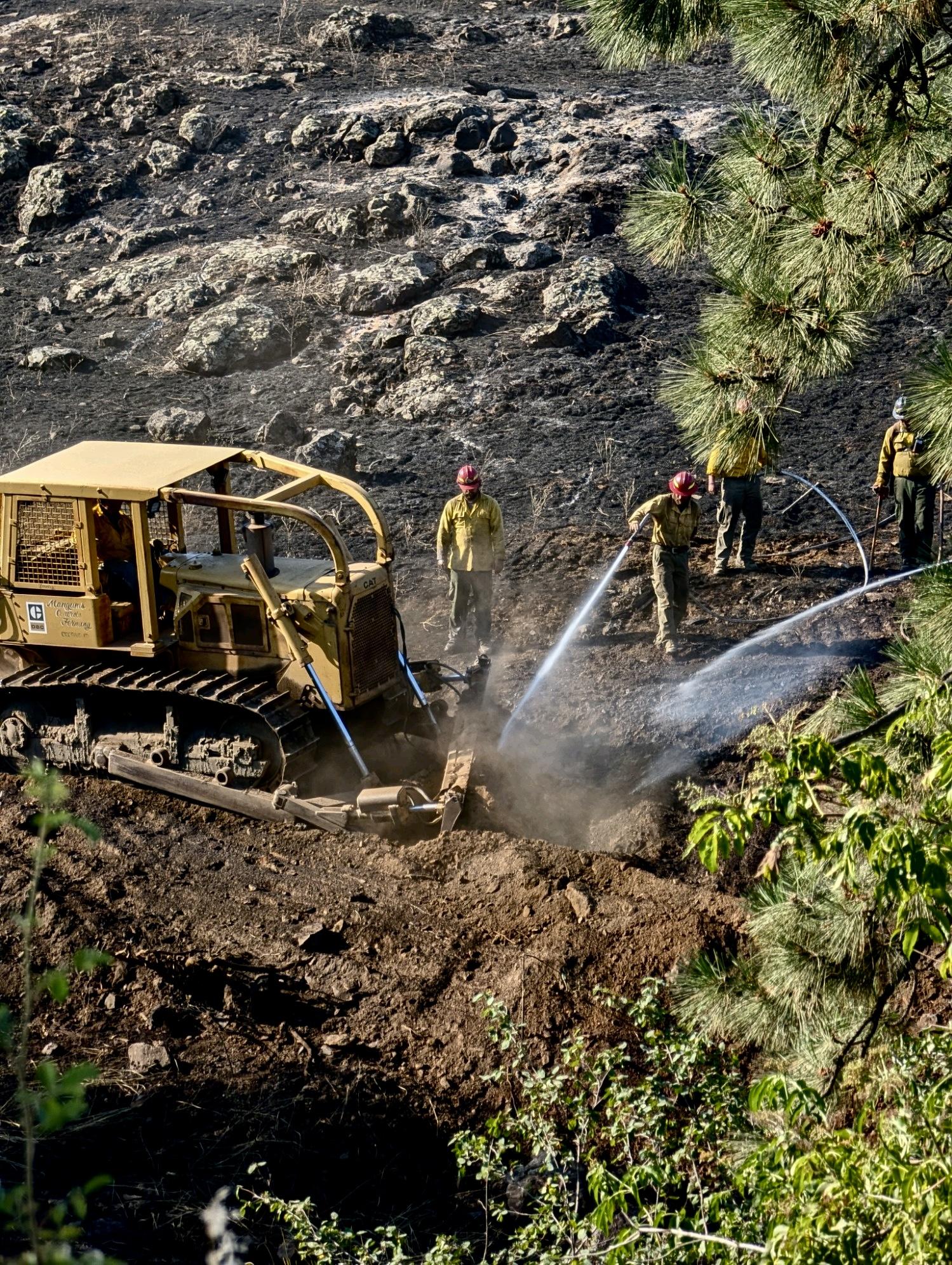 A dozer and firefighters working on a wildland fire spraying water and mixing mineral soil.