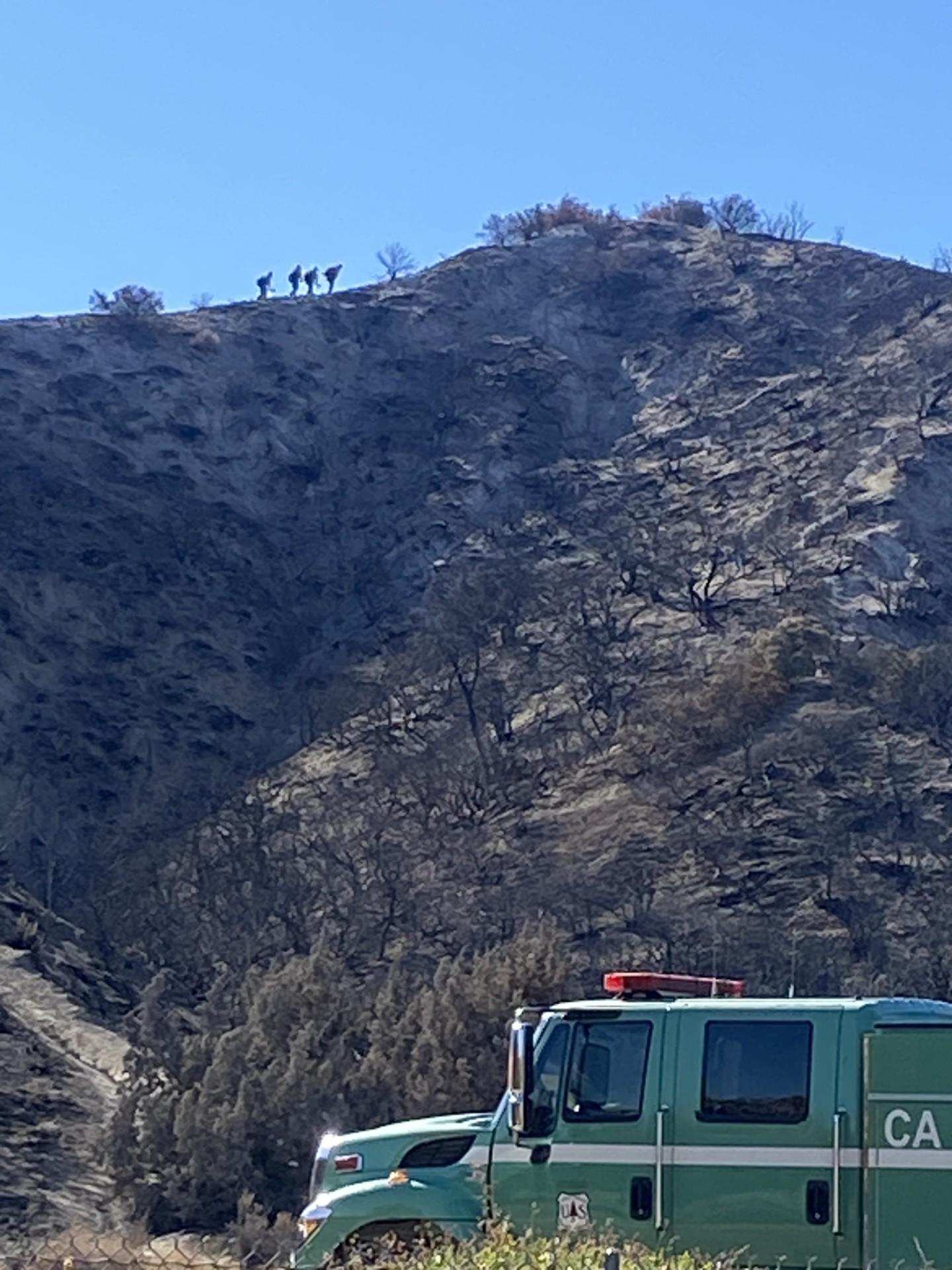 A green engine site in front of a burned hillside, with firefighters silhouetted on the ridgetop.