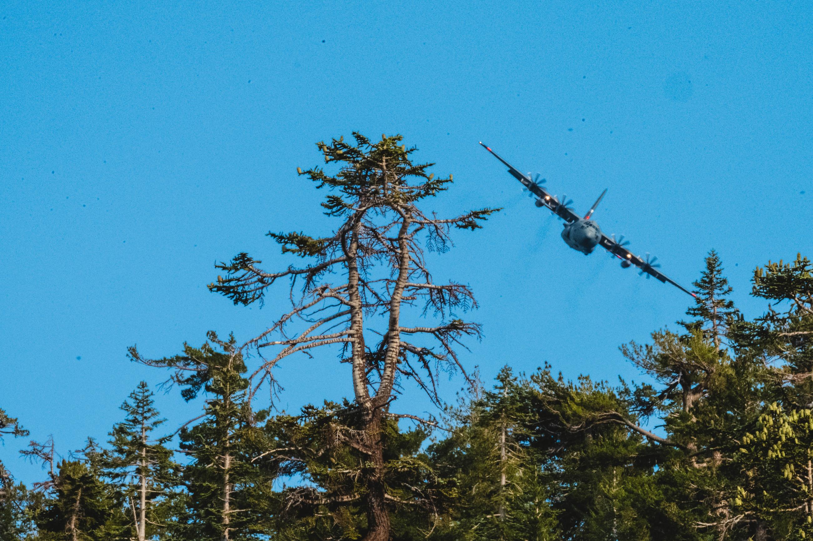 C-130 with The Modular Airborne Fire Fighting System, or MAFFS flies over green trees with blue background 