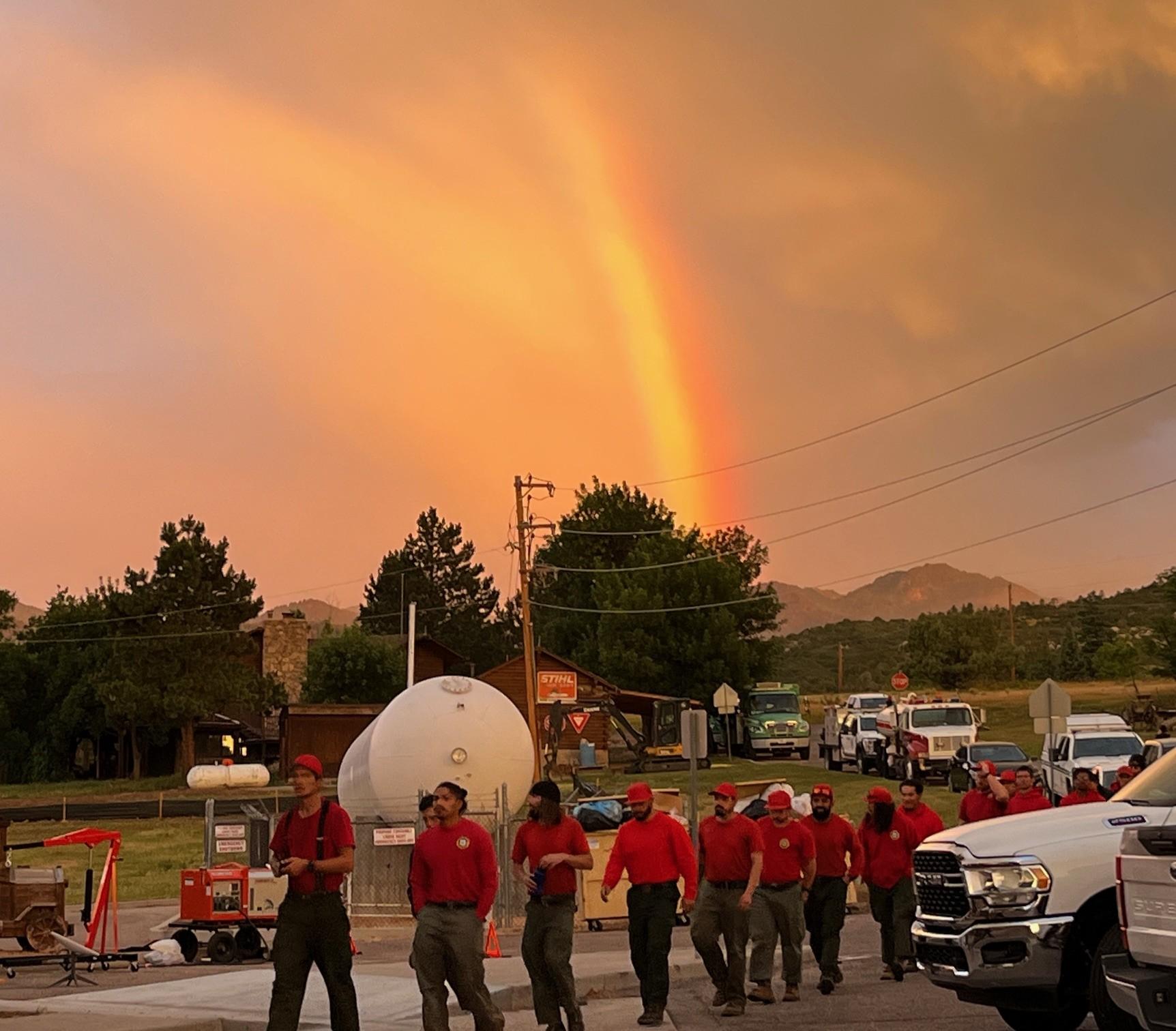 This is a cloudy shot from the morning, a rainbow is across the sky. At the bottom of the photo, several hotshots from Vista Grande Hotshots are seen walking. They are wearing red shirts and green Nomex pants.