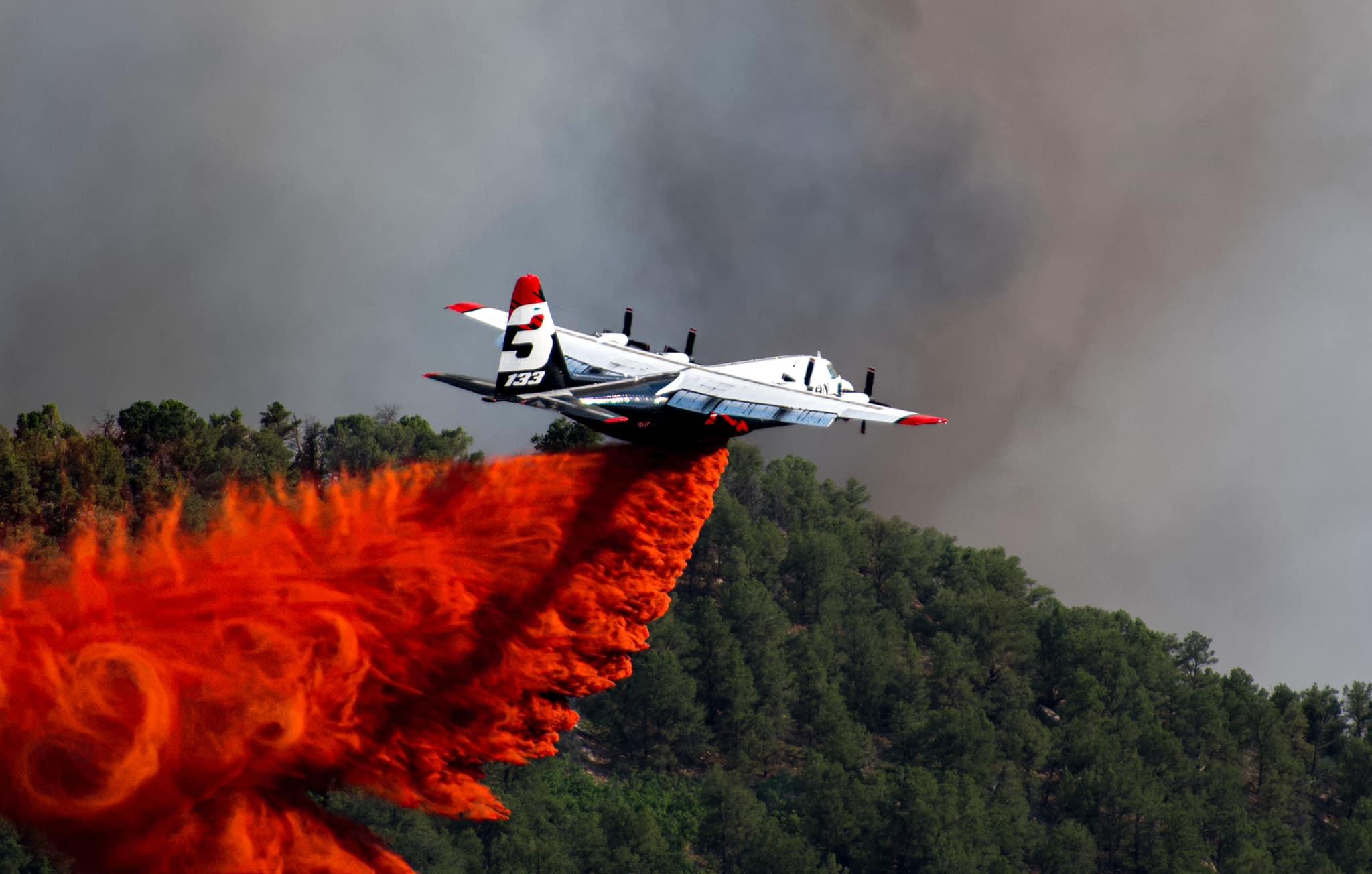 A mostly white Large Air Tanker drops bright red retardant in the foreground with green trees in the midground and black smoke in the background