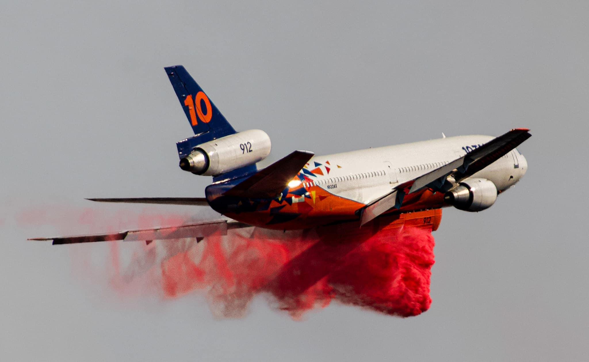 A mostly white Very Large Air Tanker (Tanker 10) drops bright red retardant in the foreground with dispersed smoke in the background