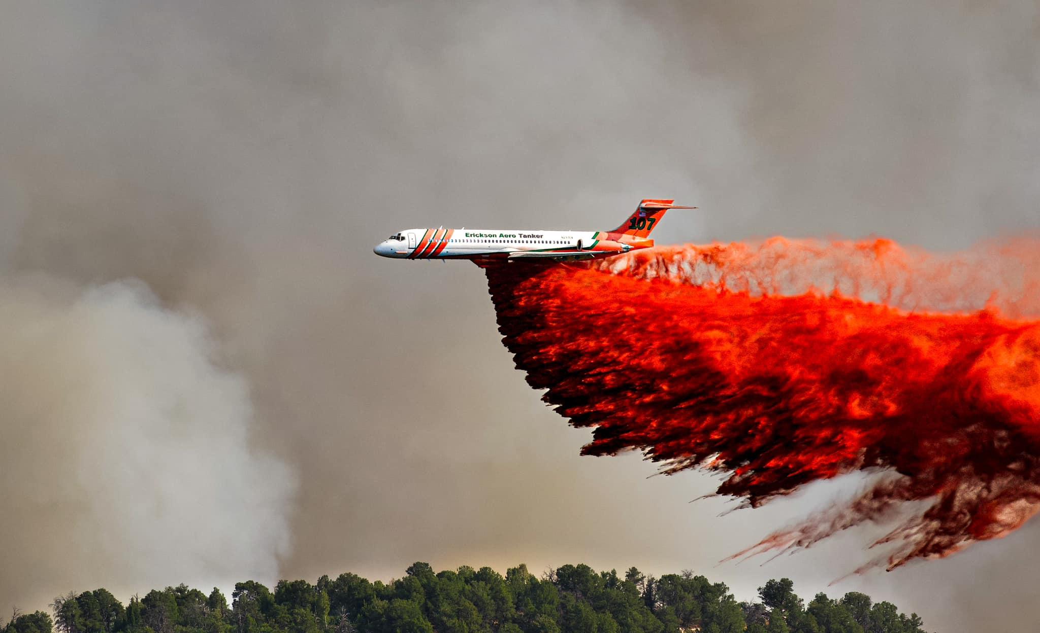 A large air tanker flies across a sky filled with black smoke dropping bright red retardant on to green trees