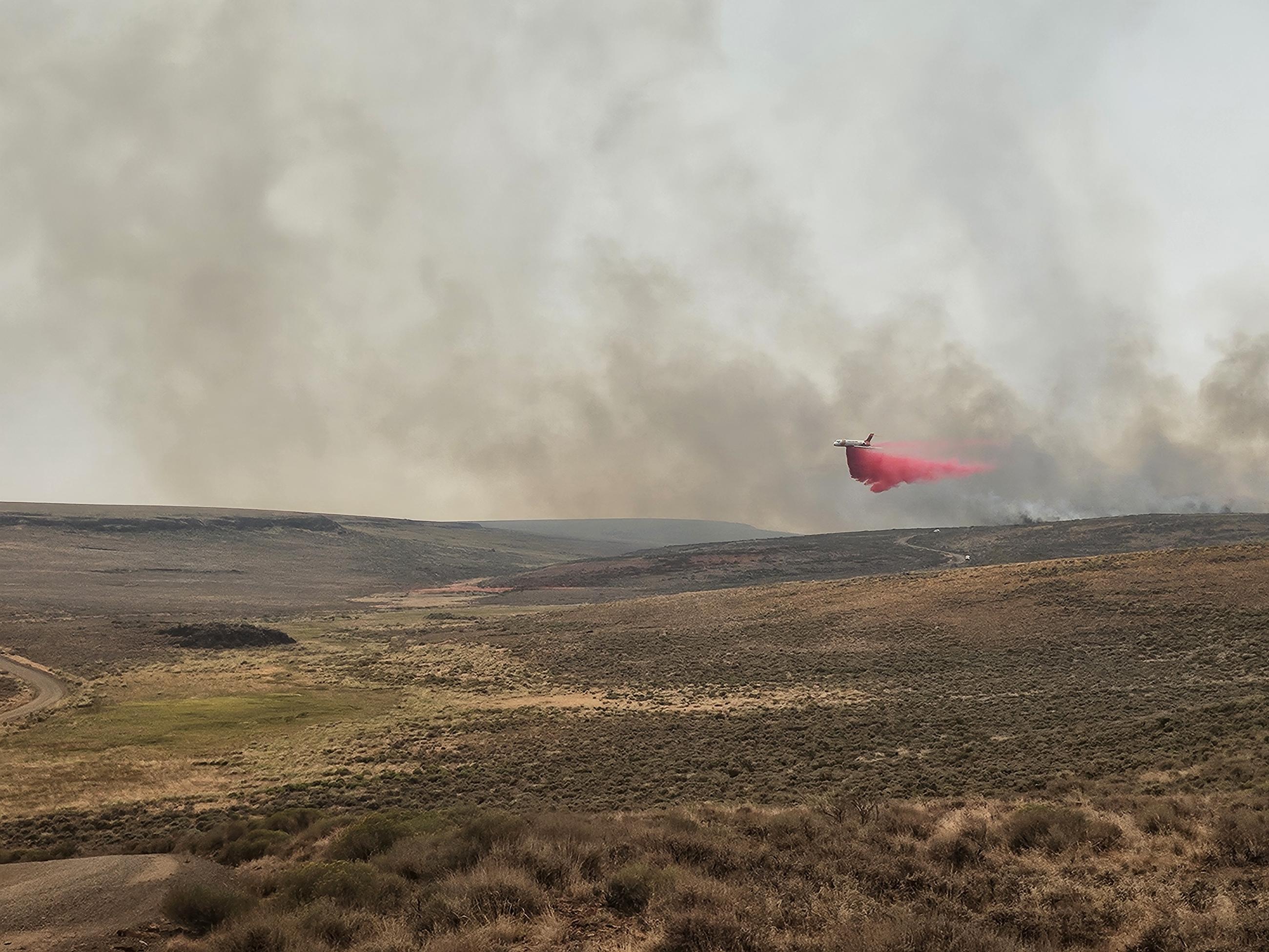 Large air tanker drops bright red retardant in advance of smoke on a moderately hilly desert landscape