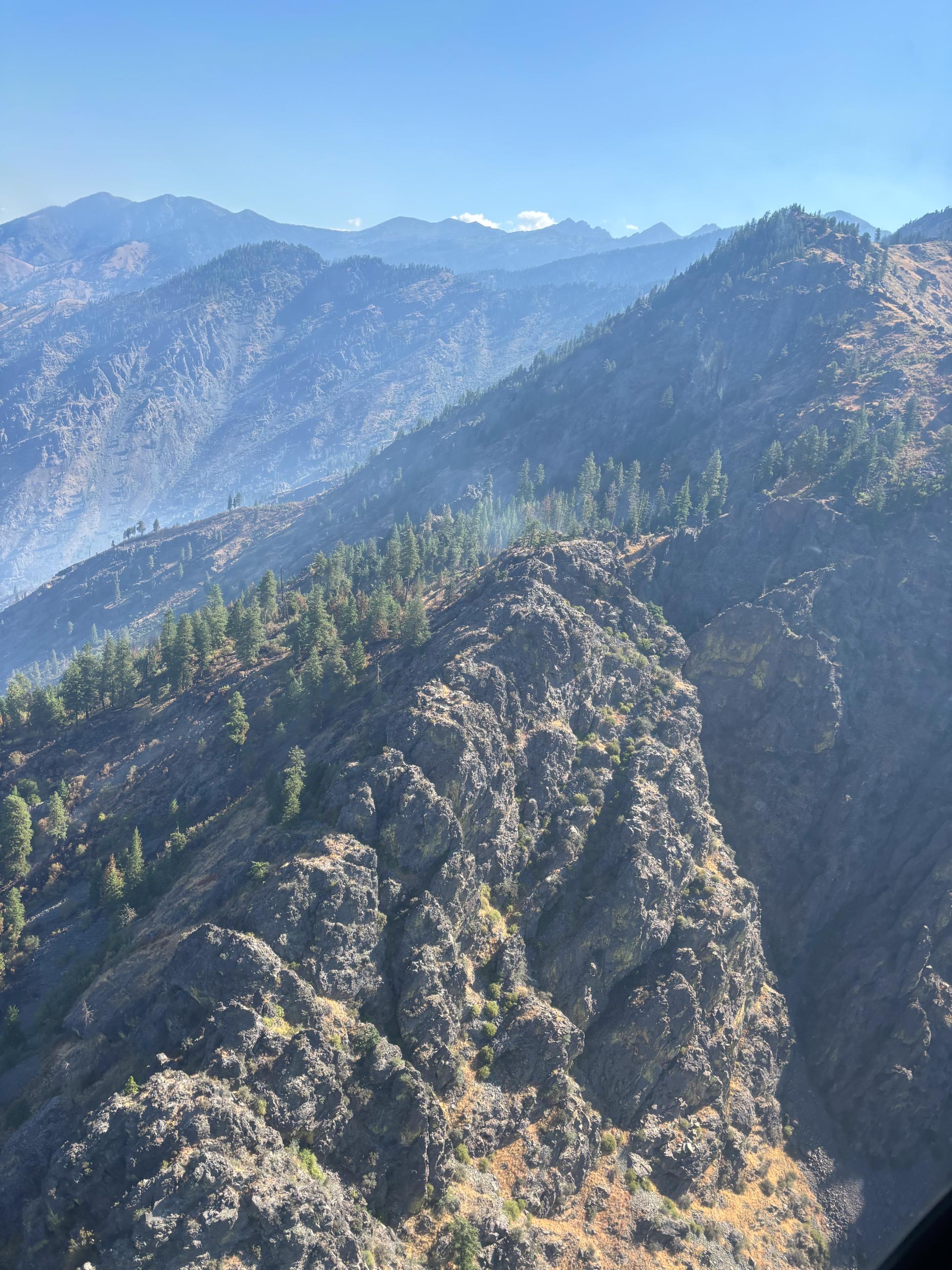 A rocky mountain with green pine trees on it. Blue sky in the background. Smoke lingers over the mountains. 