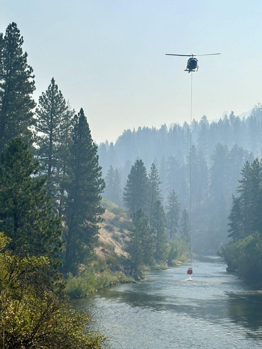 A helicopter dips a bucket into the South Fork of the Payette River to collect water to drop on the fire.