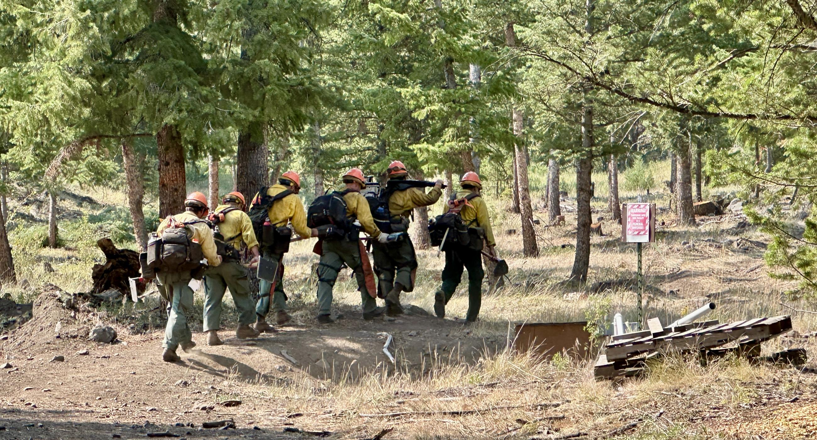 Six firefighters hike in a line, wearing fire packs and carrying tools.