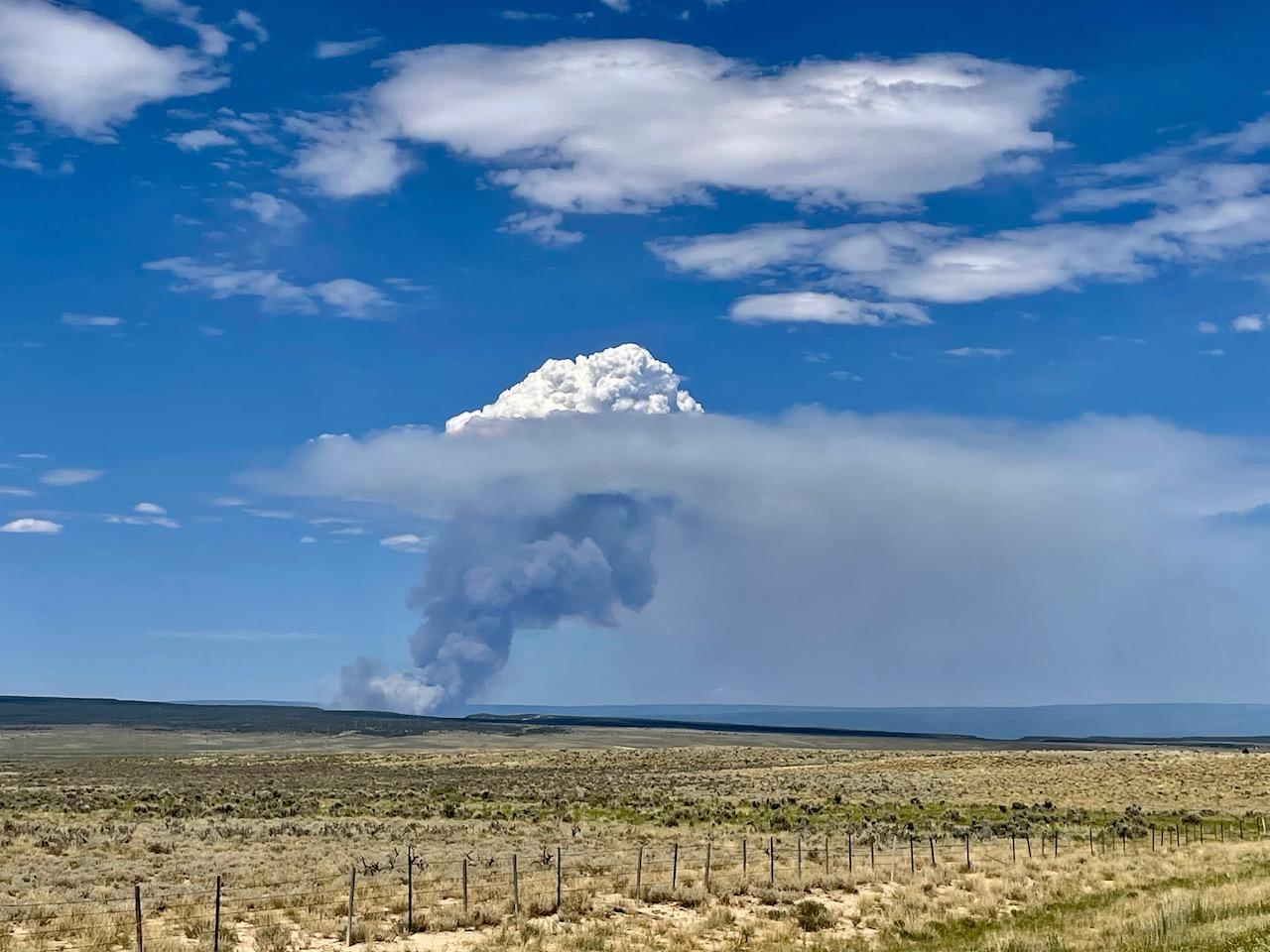 The image shows a large smoke column rising from a desert landscape