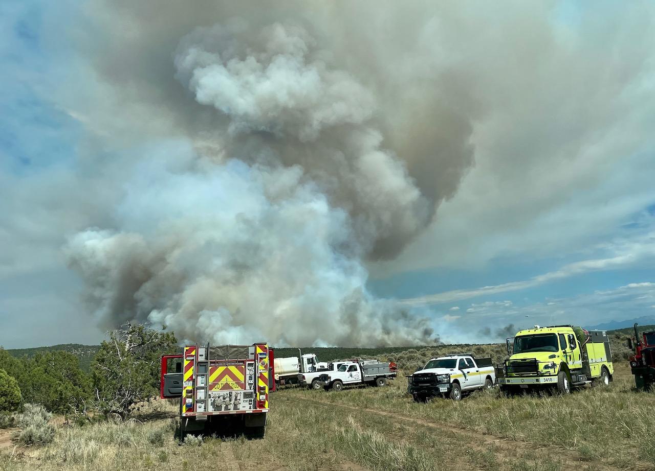 The photo shows fire engines and agency vehicles staged near the Bucktail fire with smoke rising in the background from a flat, desert landscape