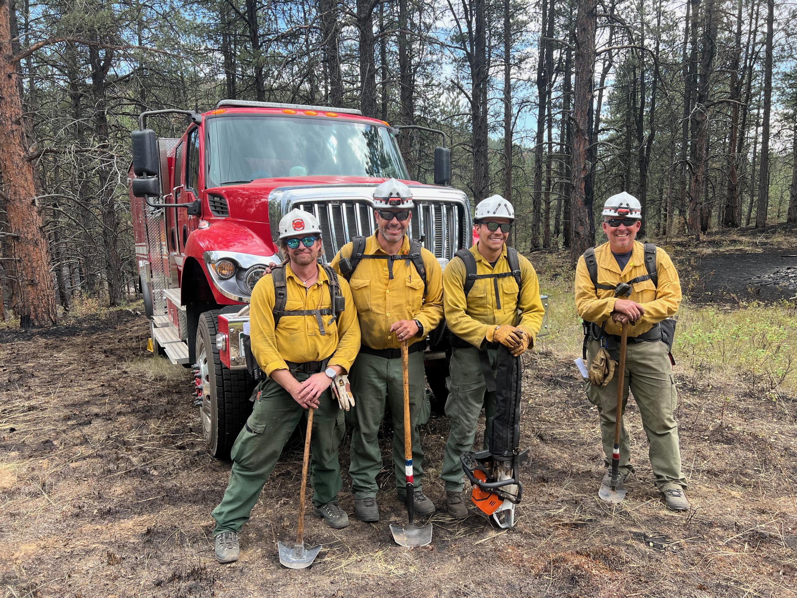 Four firefighters in yellow shirts, green fire pants, white helmets and holding fire tools are shown standing in front of their fire engine in partially burned area of national forest.