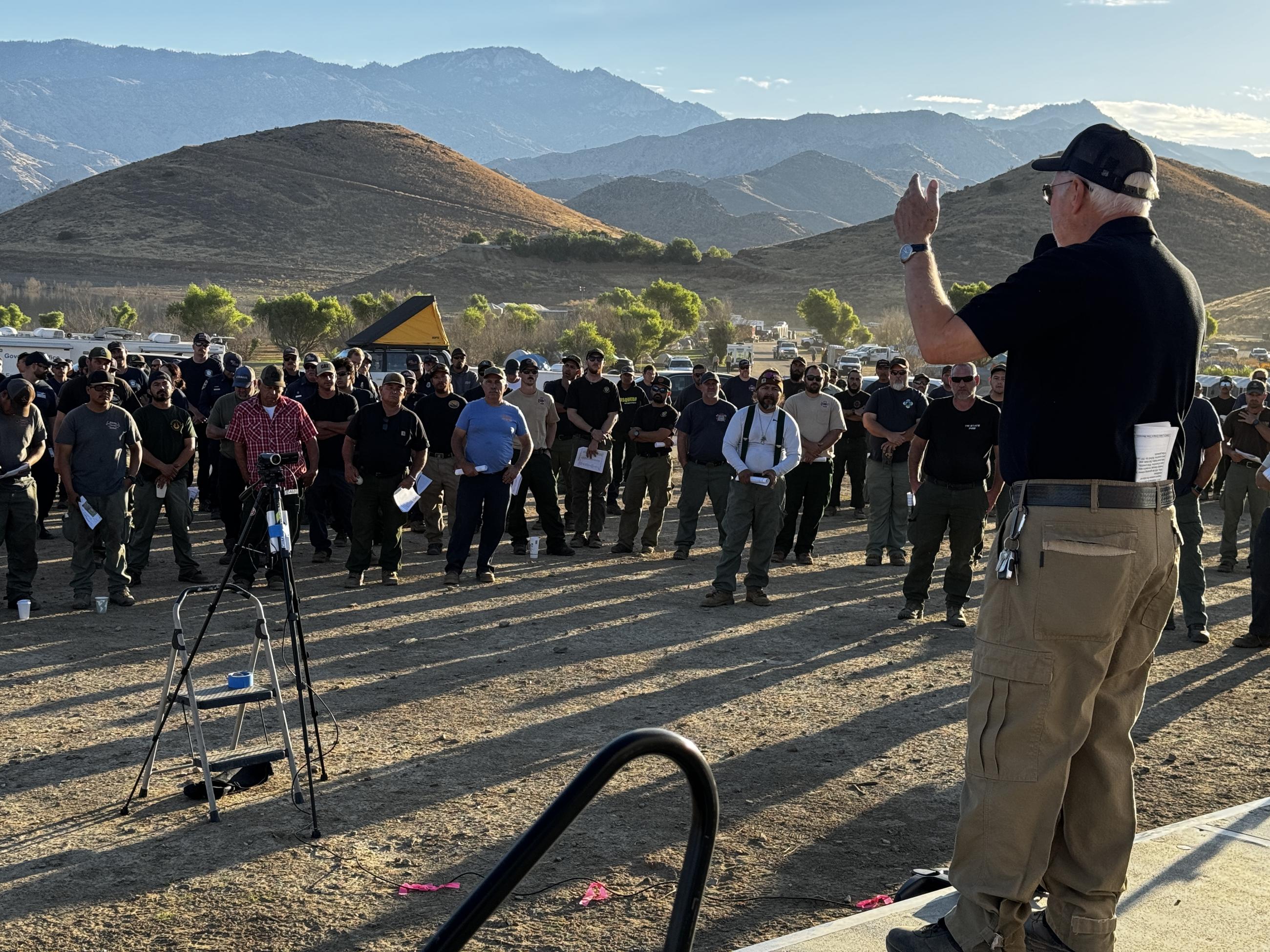 A grey-haired man in a black shirt, tan pants, and black ballcap stands on stage and speaks to a crowd of fire personnel 