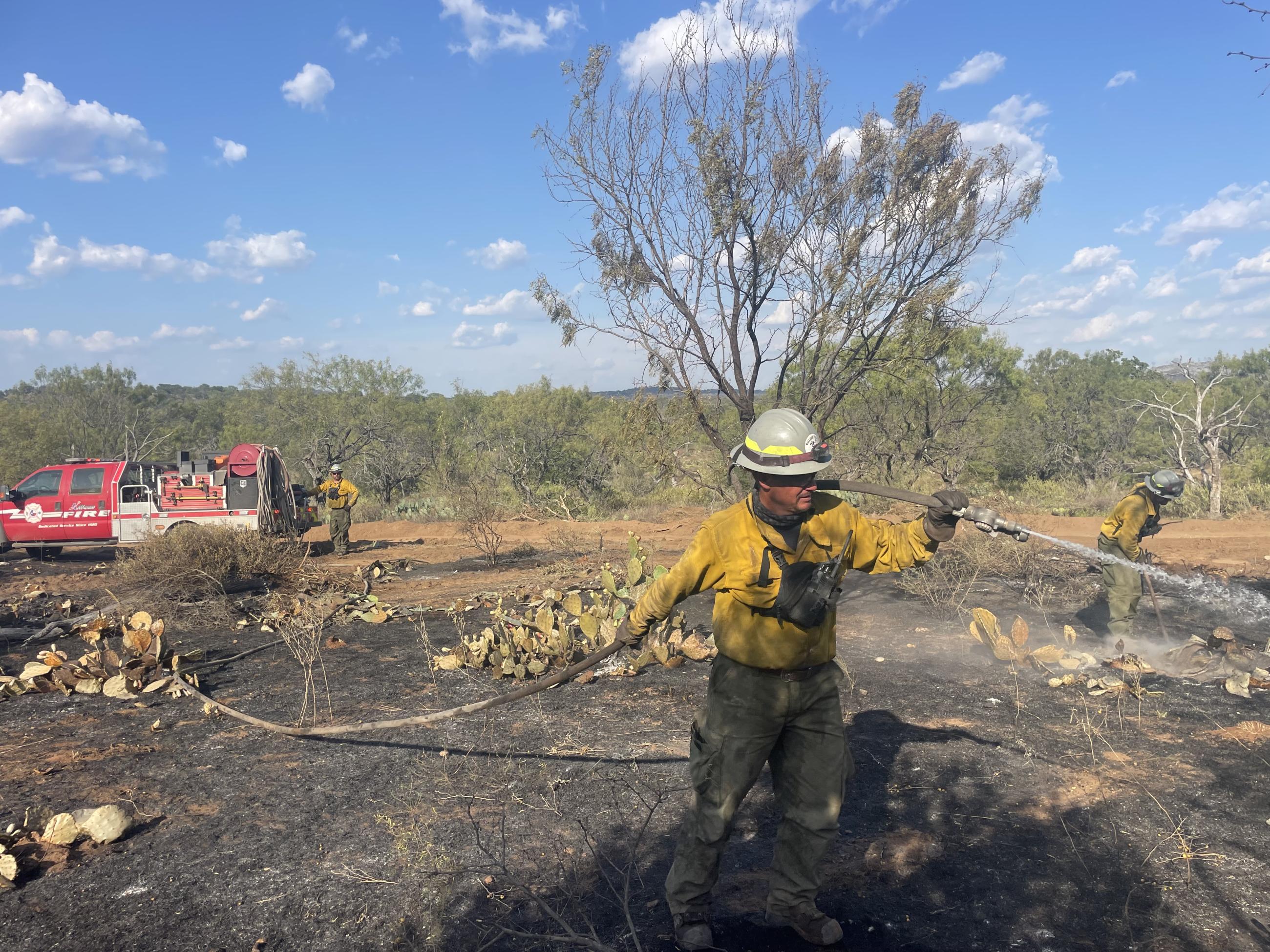 Male firefighter wearing a white hard hat, brown gloves, yellow long sleeve shirt and green pants in seen at the front of the photo holding a white firehose which is connected to a red truck or engine off to the left-hand side of the photo with another male firefighter standing at the back of the vehicle. In the background on the right-hand side of the photo is another male firefighter wearing the same color clothing and protective equipment as the male firefighter in the front of the photo.