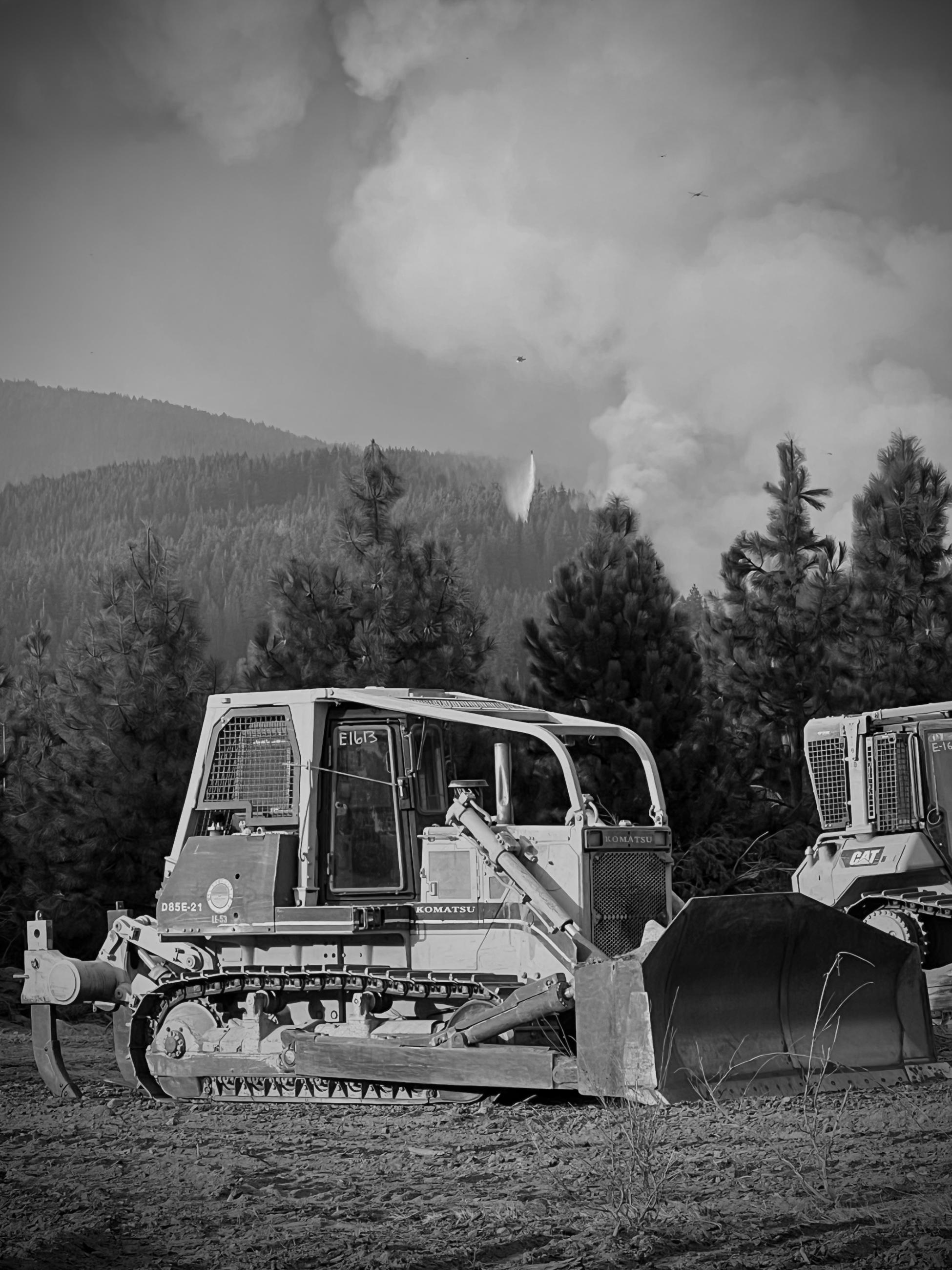 A black and white image of a bulldozer in the foreground with smoking forest in the background. A small helicopter can be seen in the distance dropping water on the fire area.