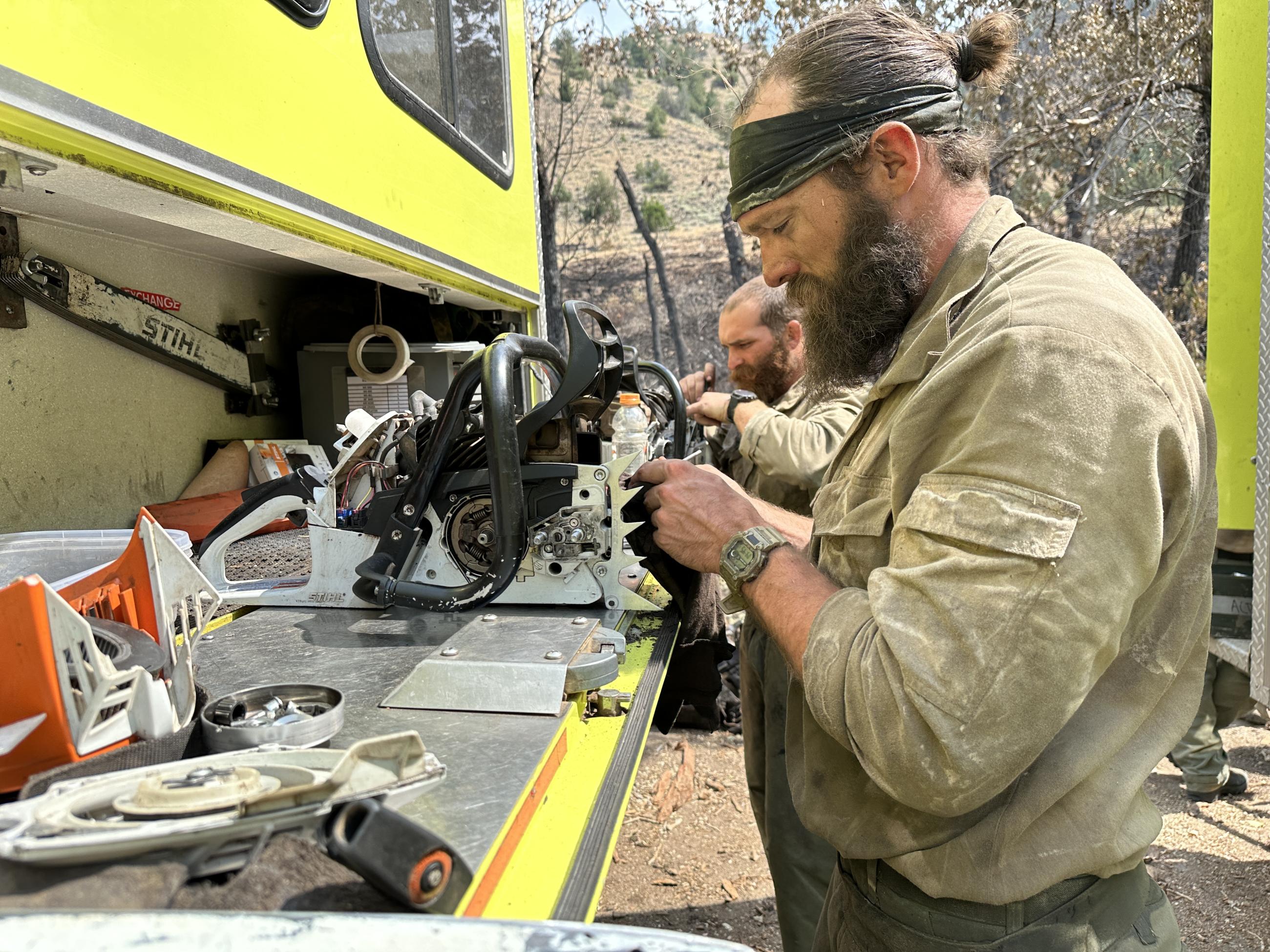 Two firefighters stand next to their truck working on their chainsaw motors