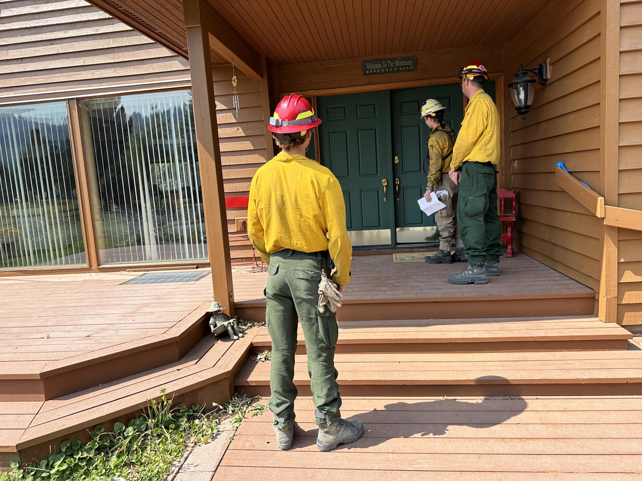 Three firefighters stand on a porch knocking on a door