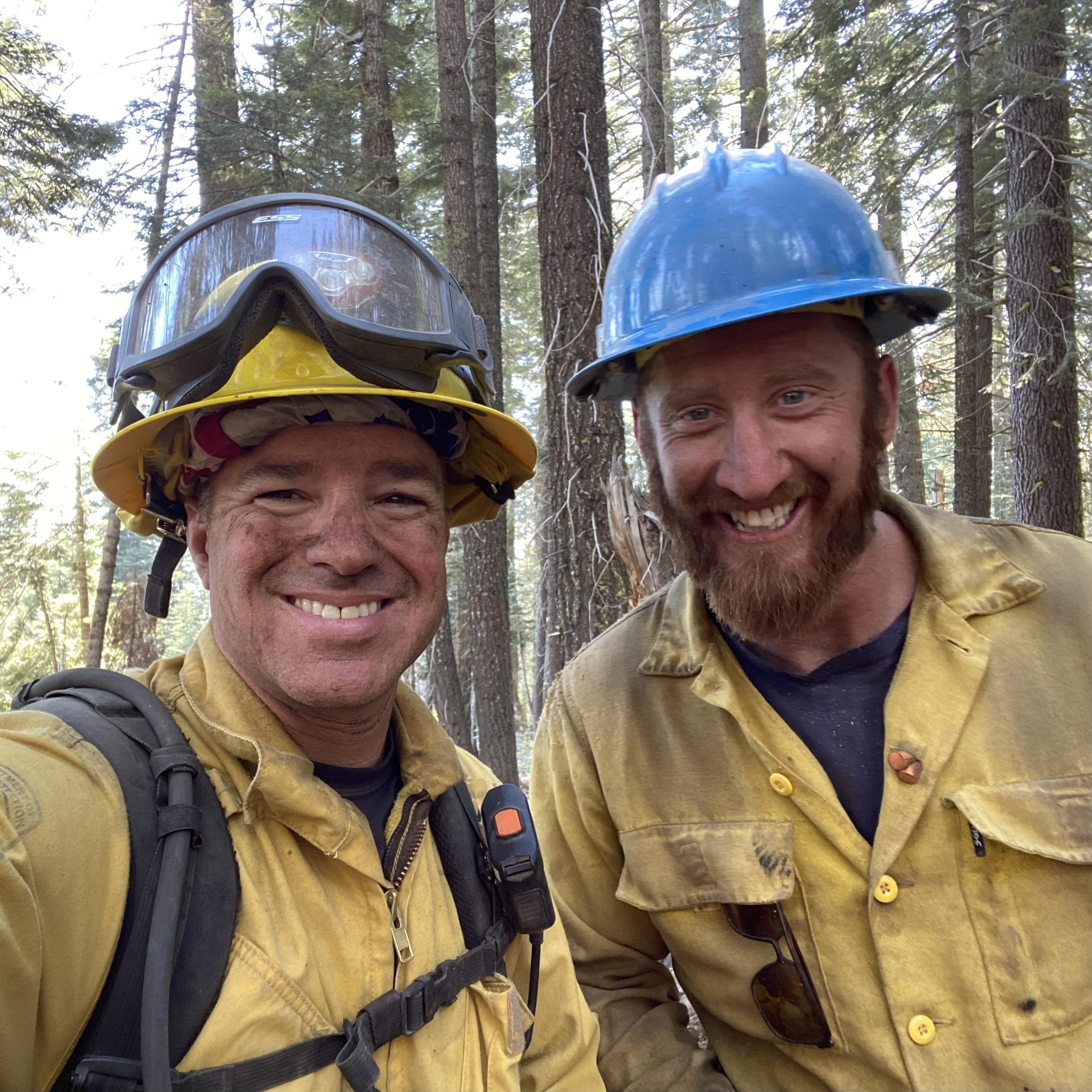 Two firefighters, one with a beard, smile at the camera. They are wearing yellow shirts and hard hats, standing in a pine forest.