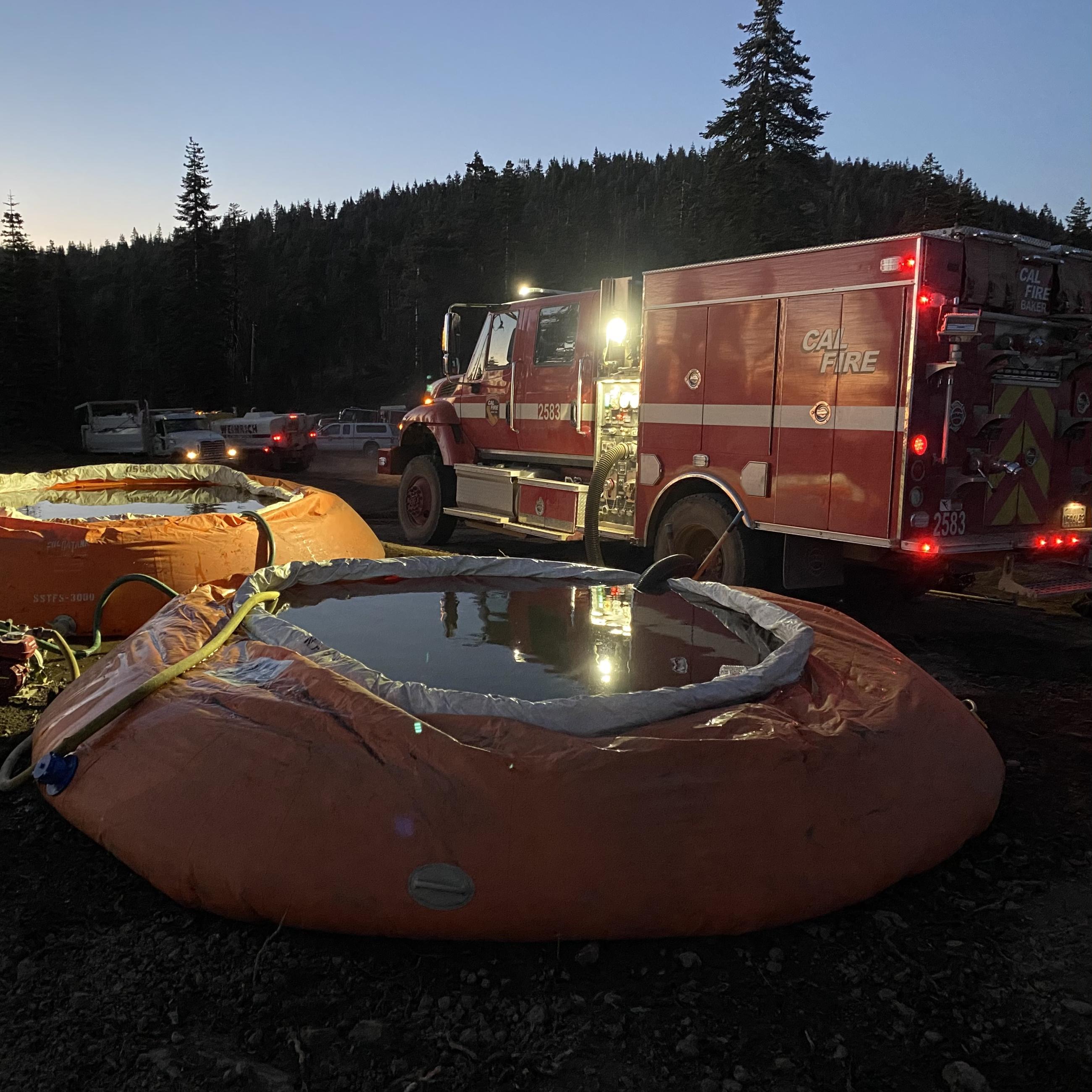 A photo taken at night just after sunset of a red water tender fire truck beside two bright orange portable water tanks with hoses and pumps