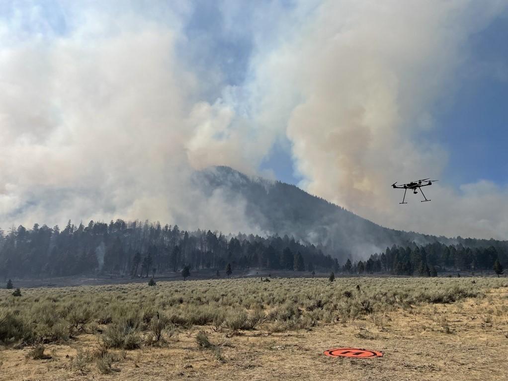 A drone flies with smoke in the background. Leaving landing pad in the sagebrush and headed to the forested area.