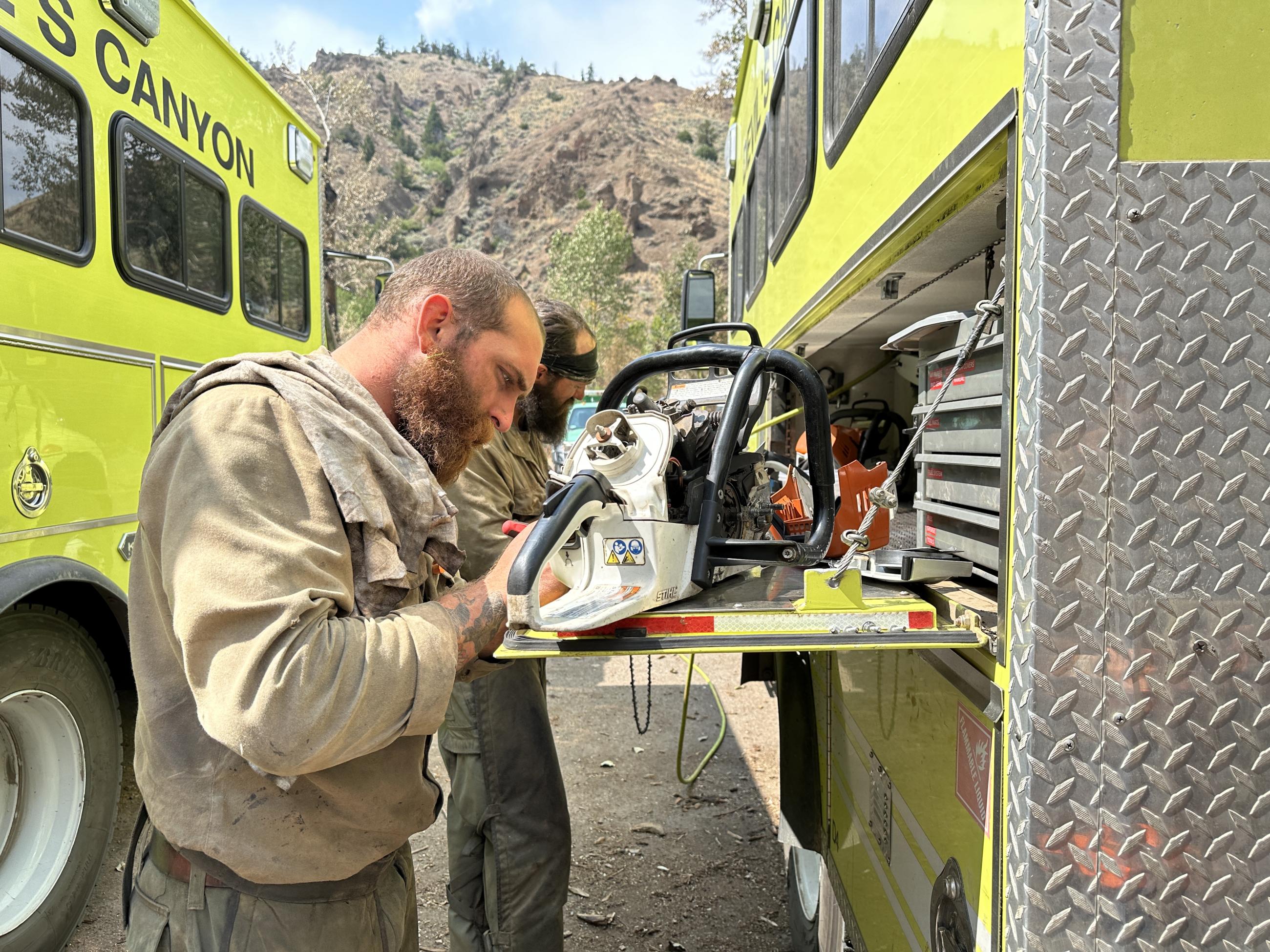 A firefighter with soot on his face works on a chainsaw next to his truck