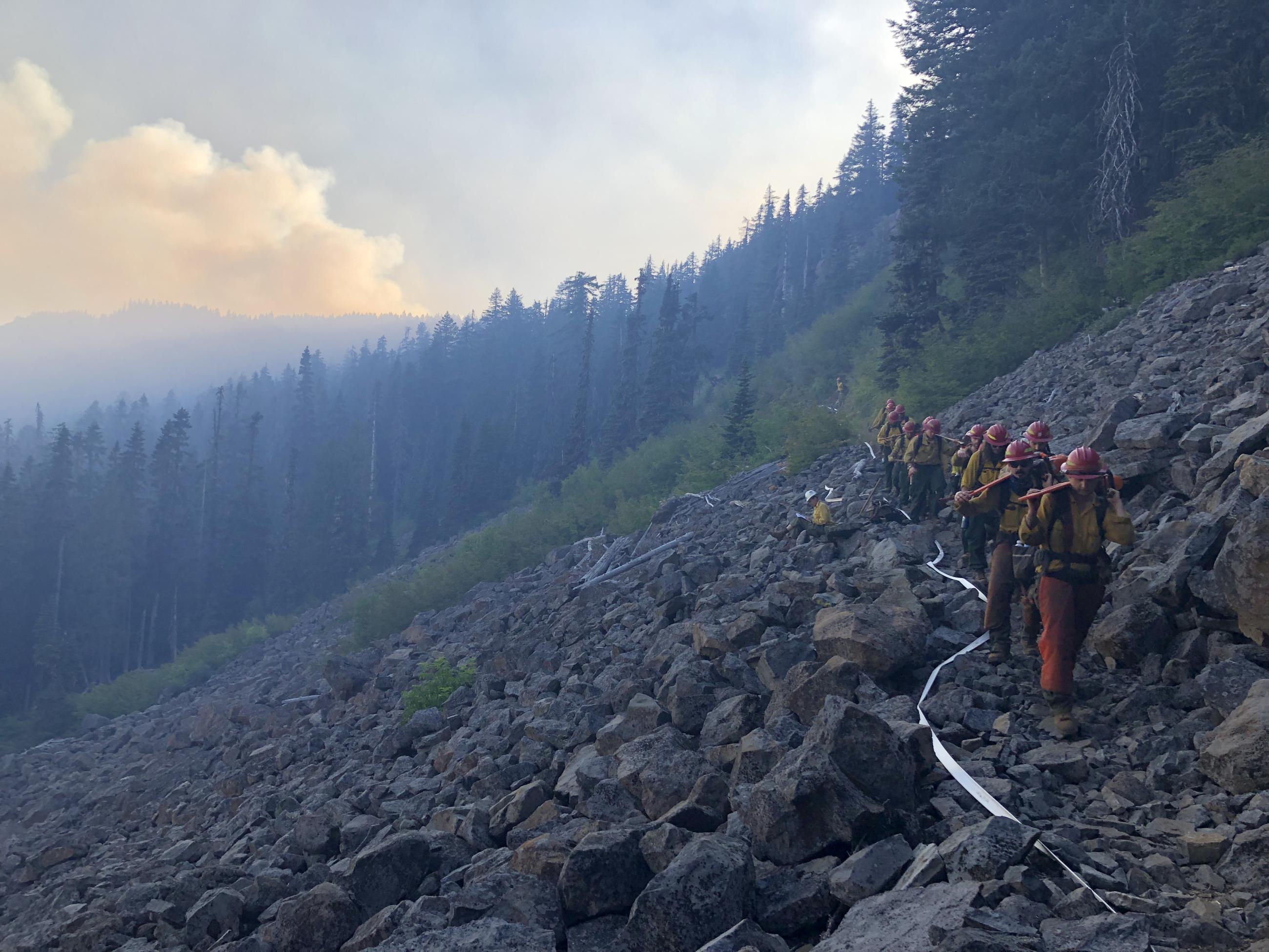 A line of firefighters with tools hike towards the viewer over large black rocks with a smoky forest in the background