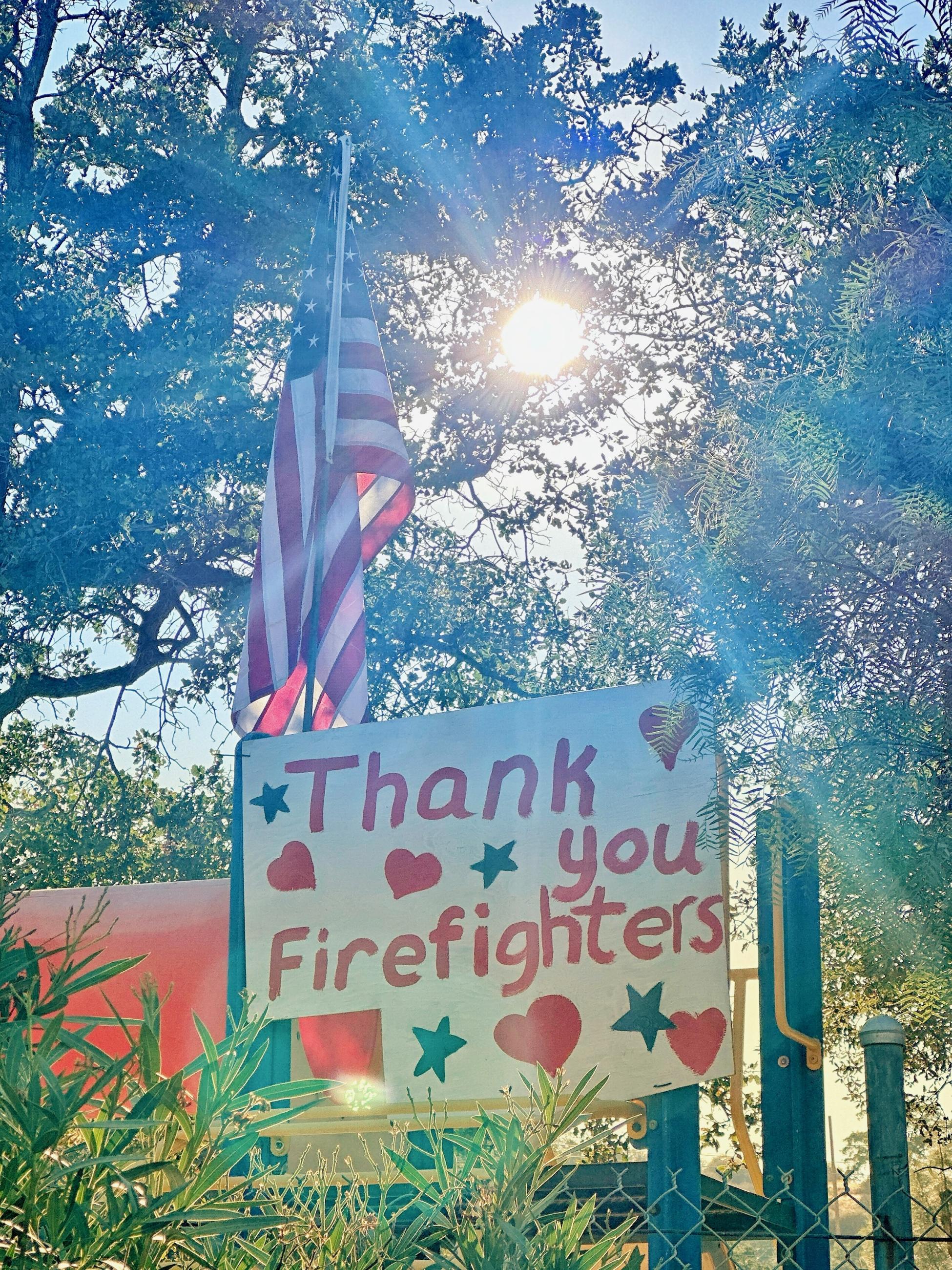 A homemade thank you sign rests next to an American flag in front of trees with the sun shining through