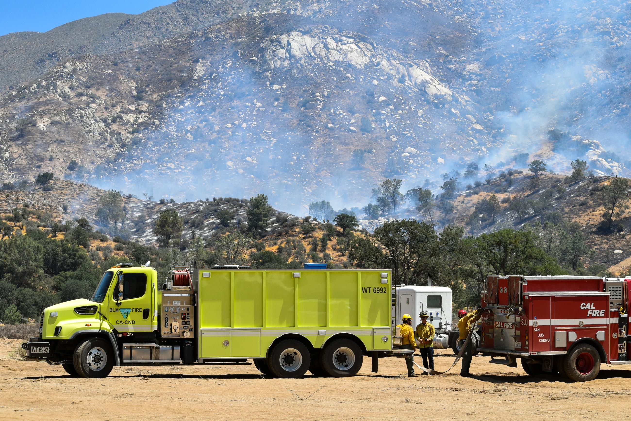 A bright yellow BLM water tender truck provides water to a red CalFire engine with a white flatbed semi in the background. The land behind the vehicles smokes