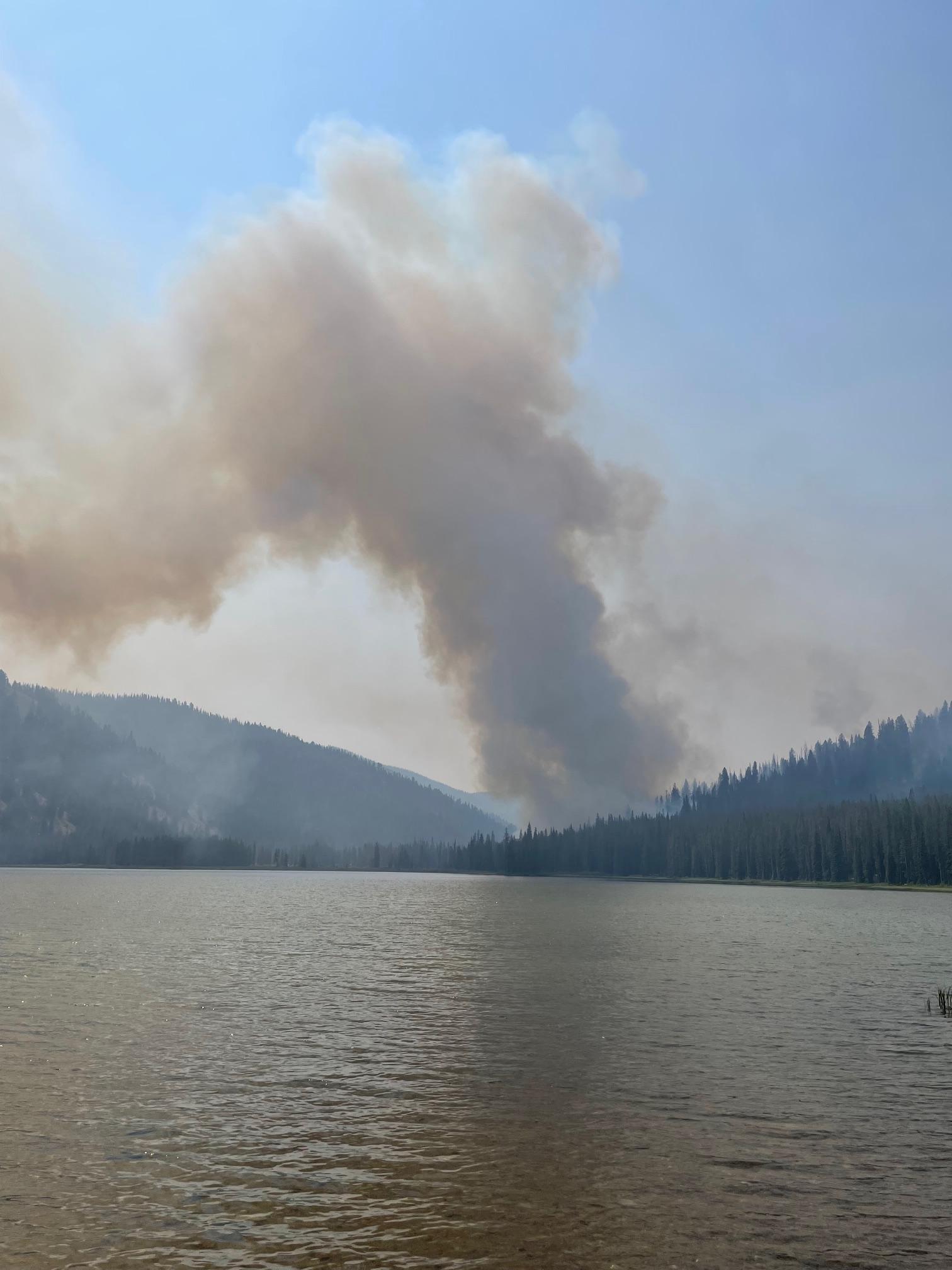 Image displays smoke rising in the distance with Bulltrout Lake in the foreground.