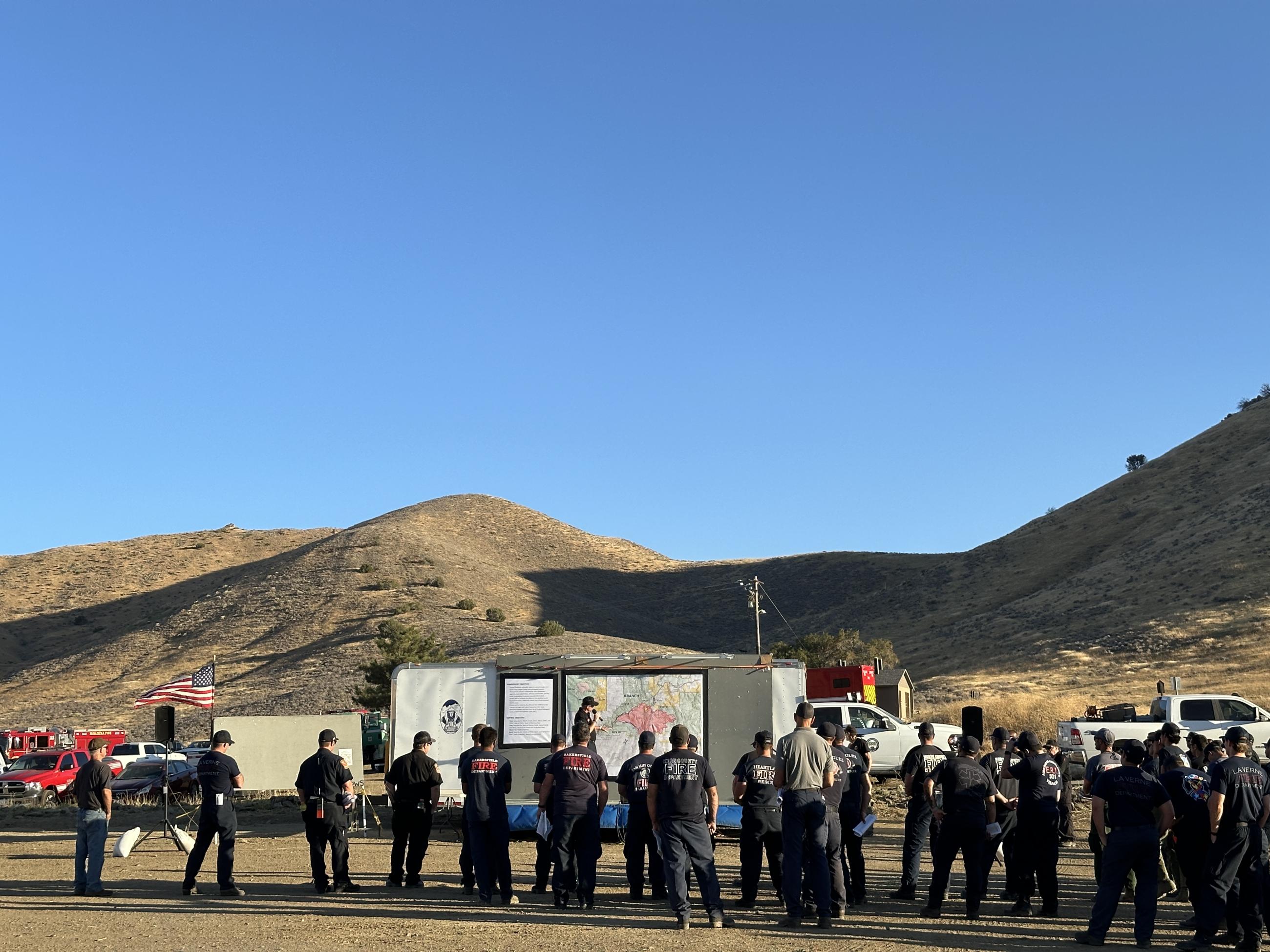 Firefighters stand in a line in front of a large map on a sunny day