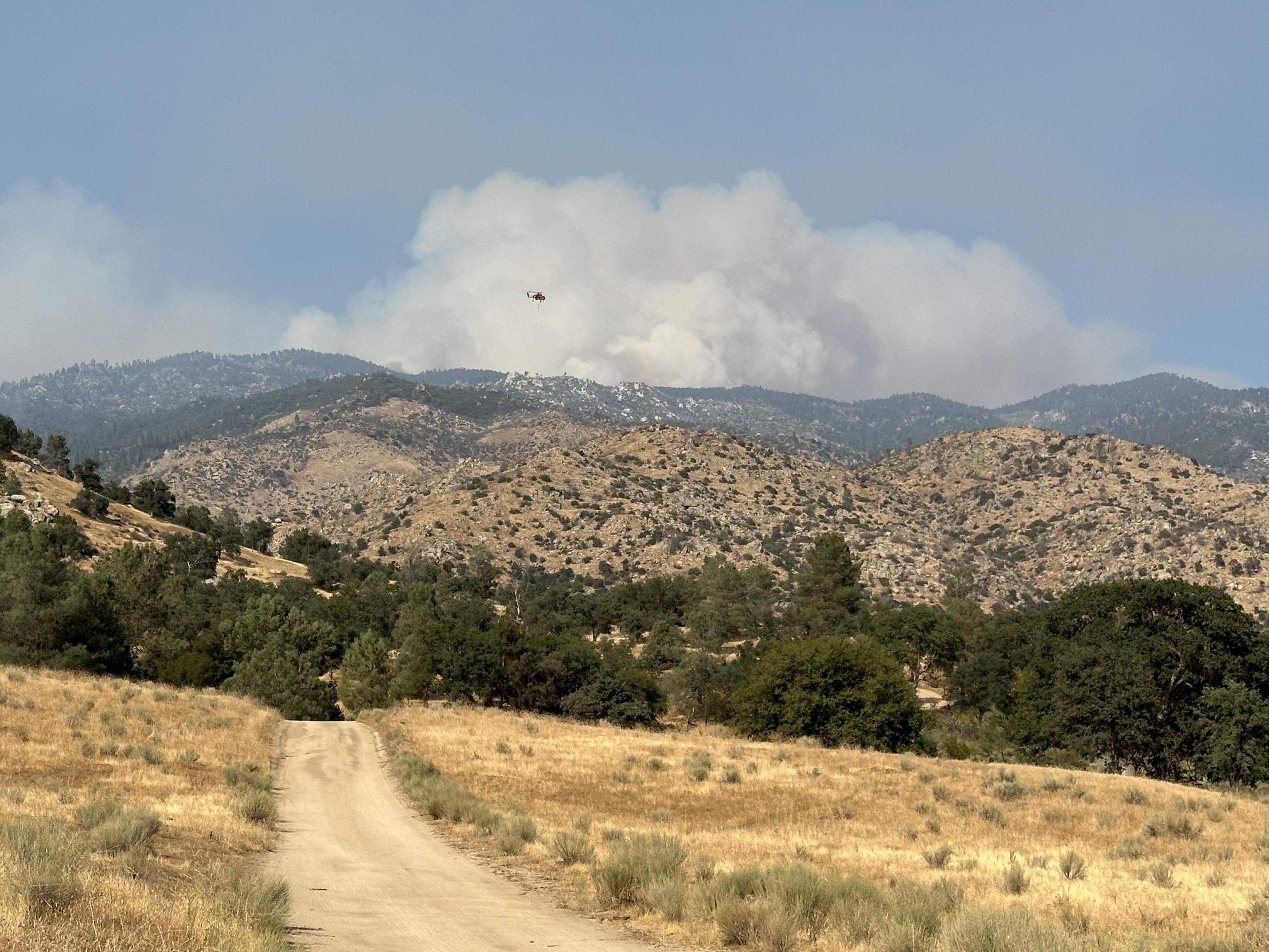 A dirt road leads into rolling hillsides with a large smoke cloud and helicopter