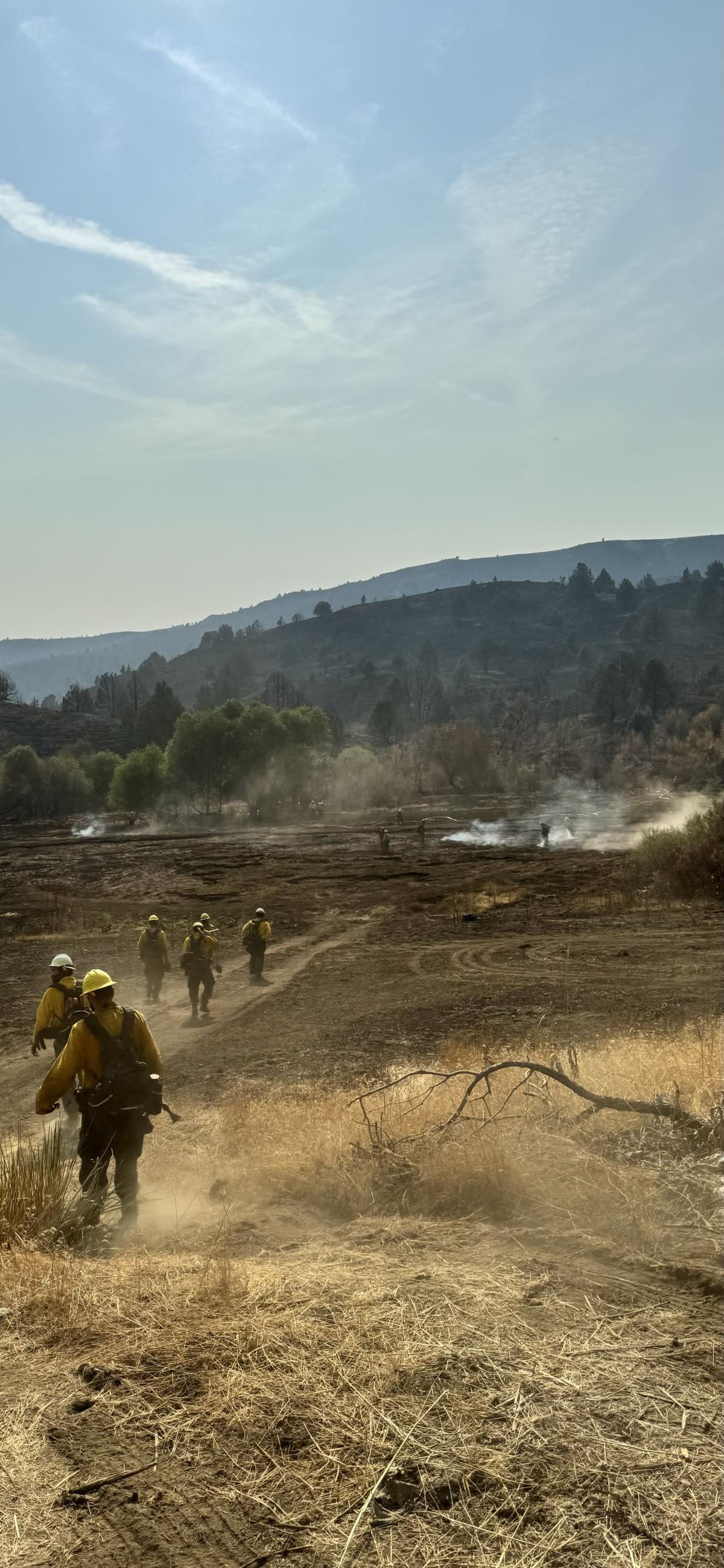 A line of firefighters in personal protective equipment walk down a dirt road towards a blackened area with smoke rising and green hillsides in the distance