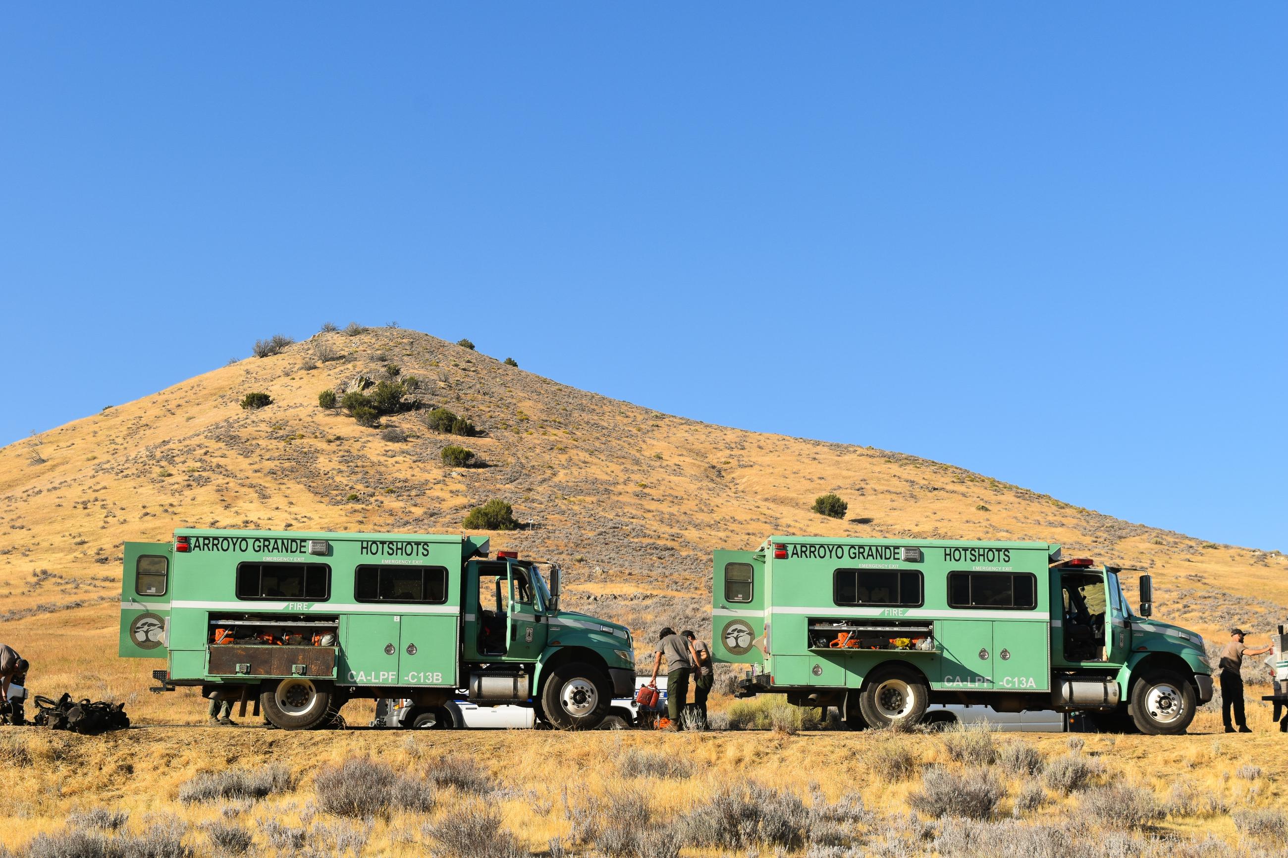 Two green hotshot buggies are parked and firefighters wearing green pants and grey tee shirts unpack fuel cans, fire packs, and other gear in a desert landscape