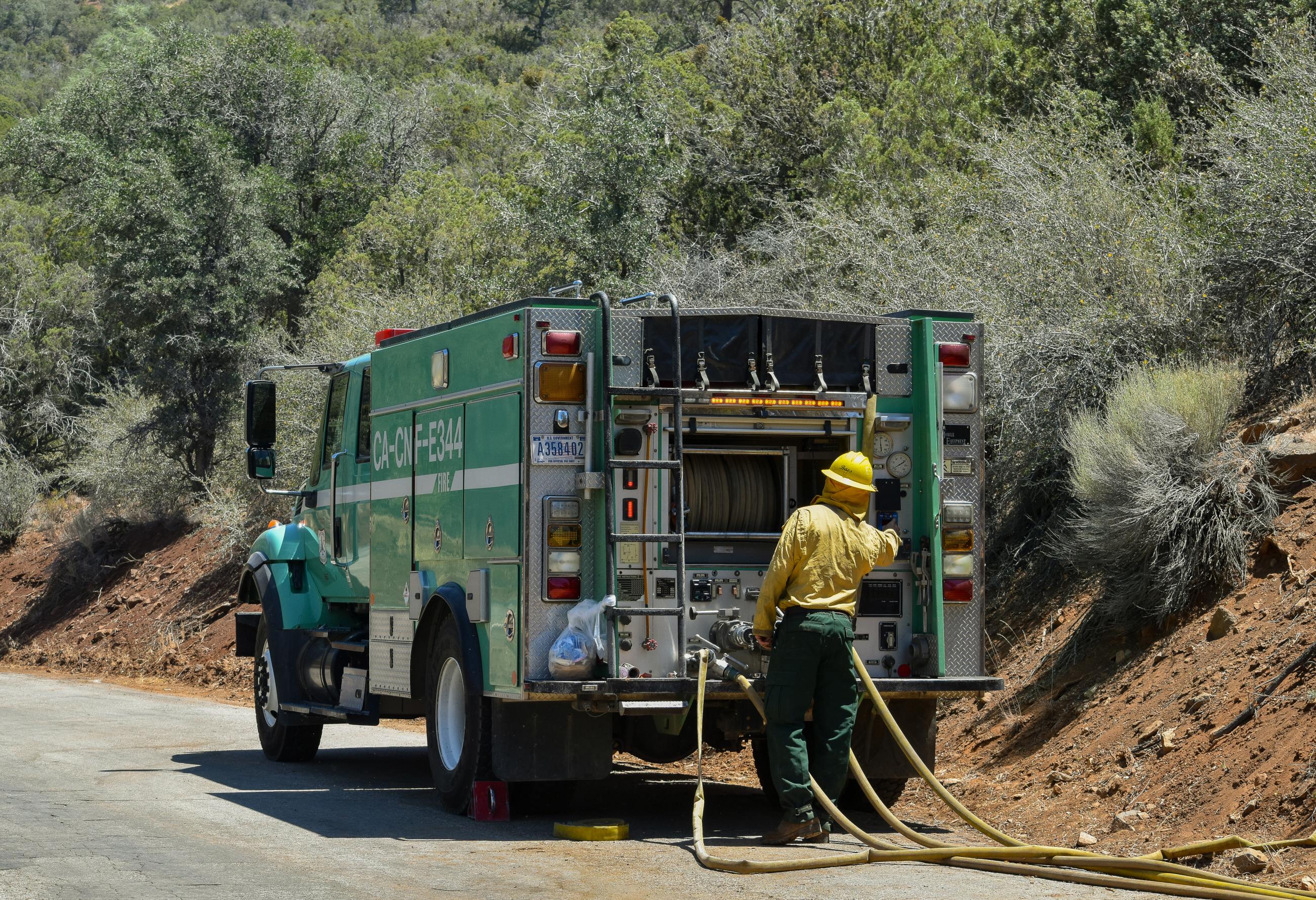 A firefighter wearing green pants, a yellow shirt, and a yellow hard hat enginners off the back of a green fore service fire truck with yellow fire hose flaked out behind it.