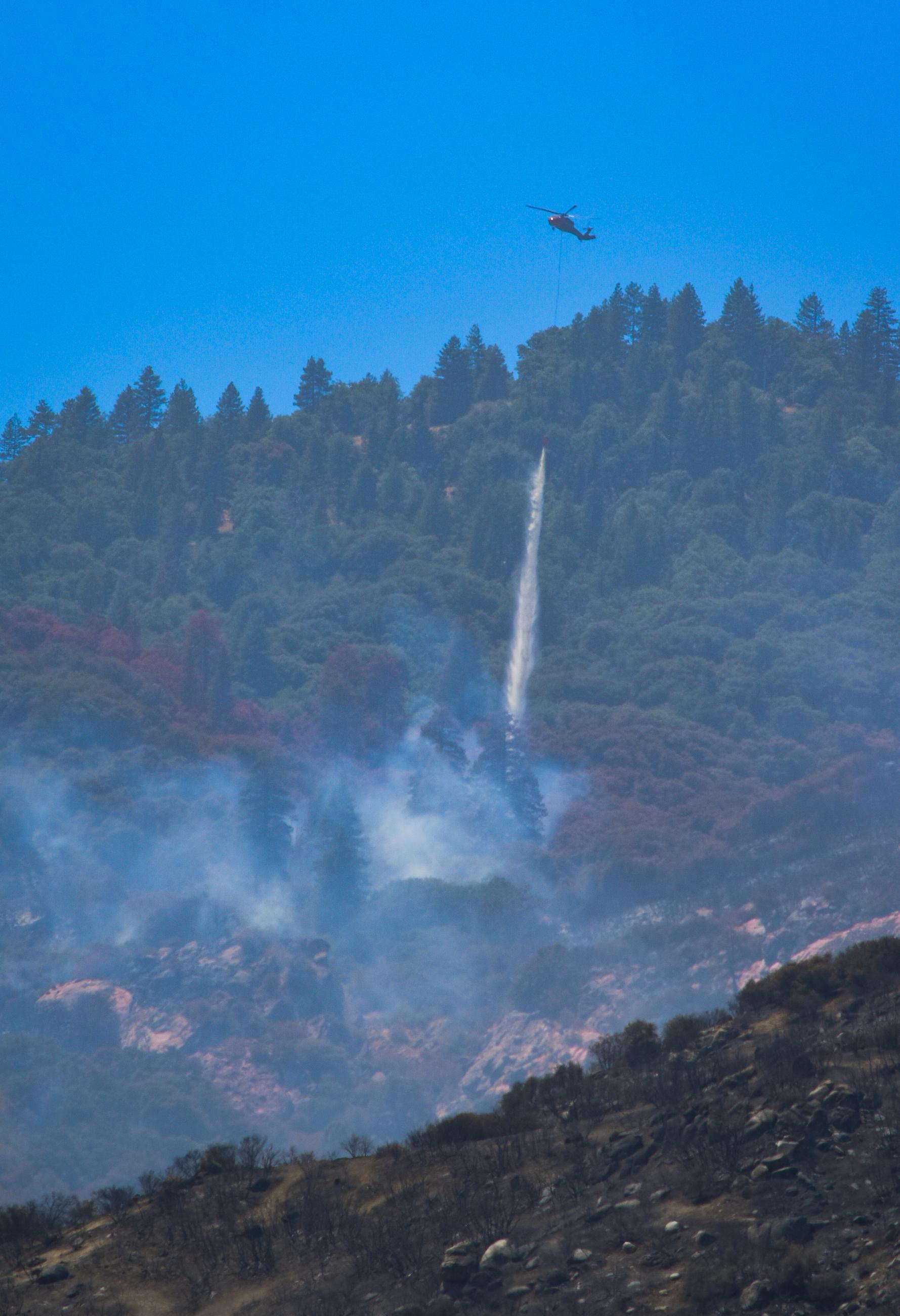 A red helicopter drops water into a forest emitting smoke, with red retardant line visible on nearby trees