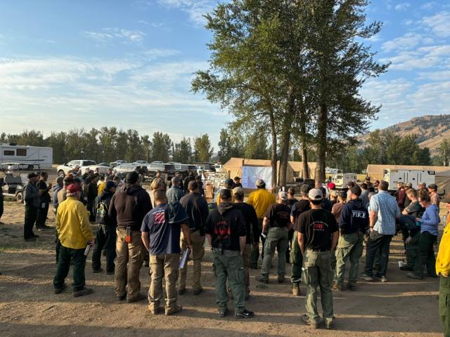 A group of firefighters stands around a board and a speaker, receiving a briefing. They are surrounded by trucks and tents at base camp.