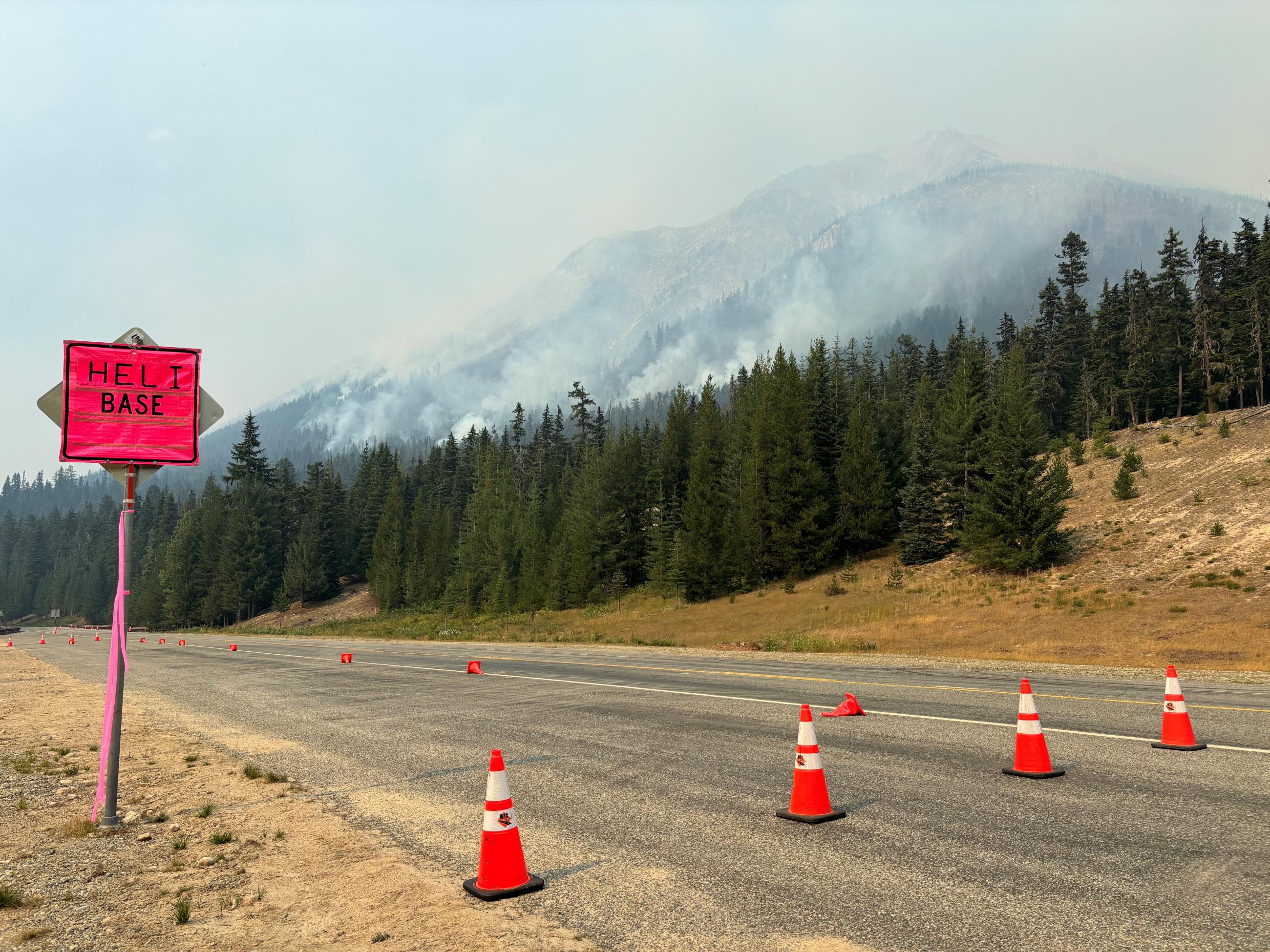 The foreground is a sign reading "helibase" with cones blocking one lane of the two lane highway, the background is a steep, timbered mountain smoldering 