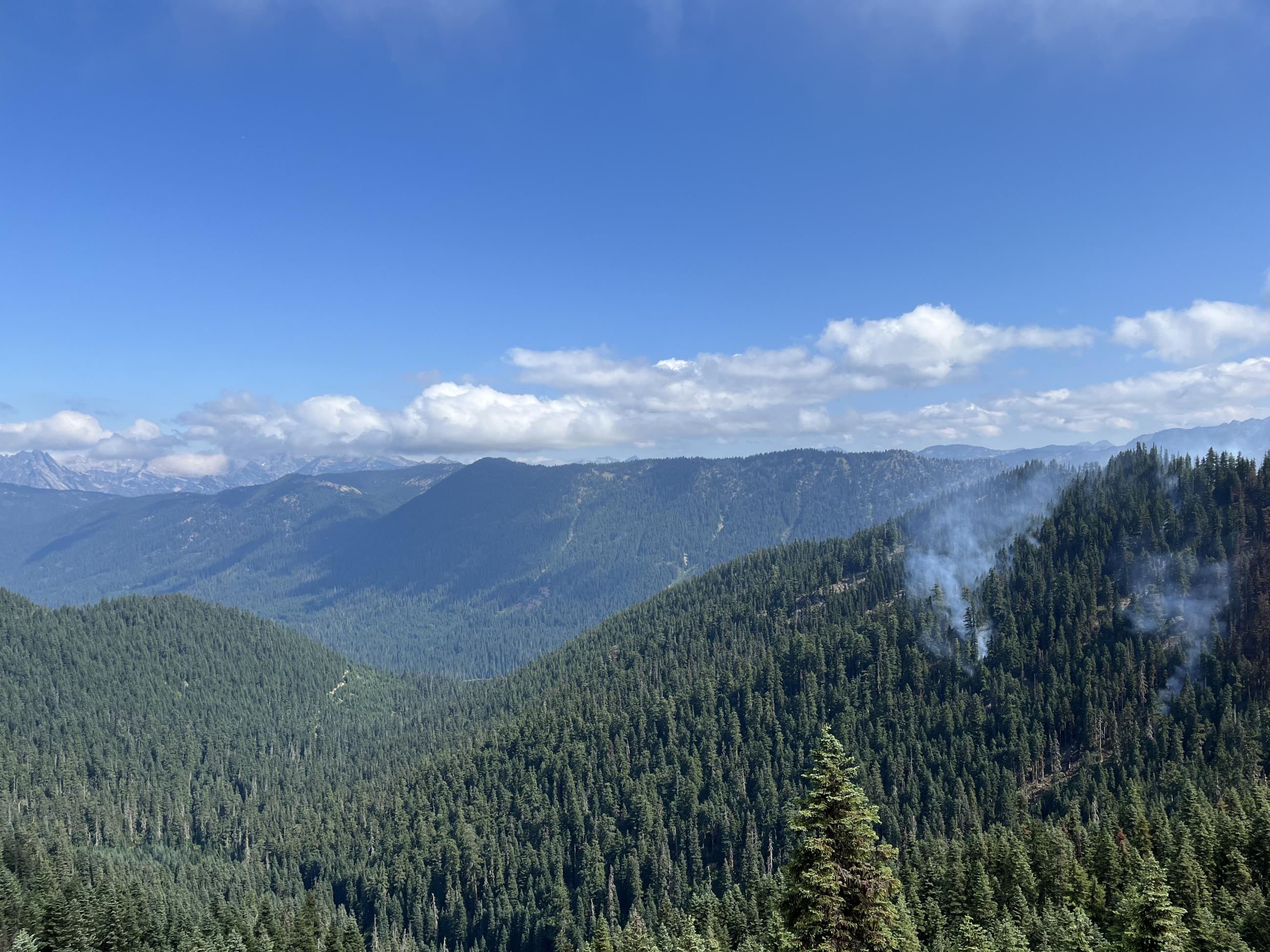View from high ridge overlooking mountains covered in coniferous forest with smoke rising from the ridge to the right