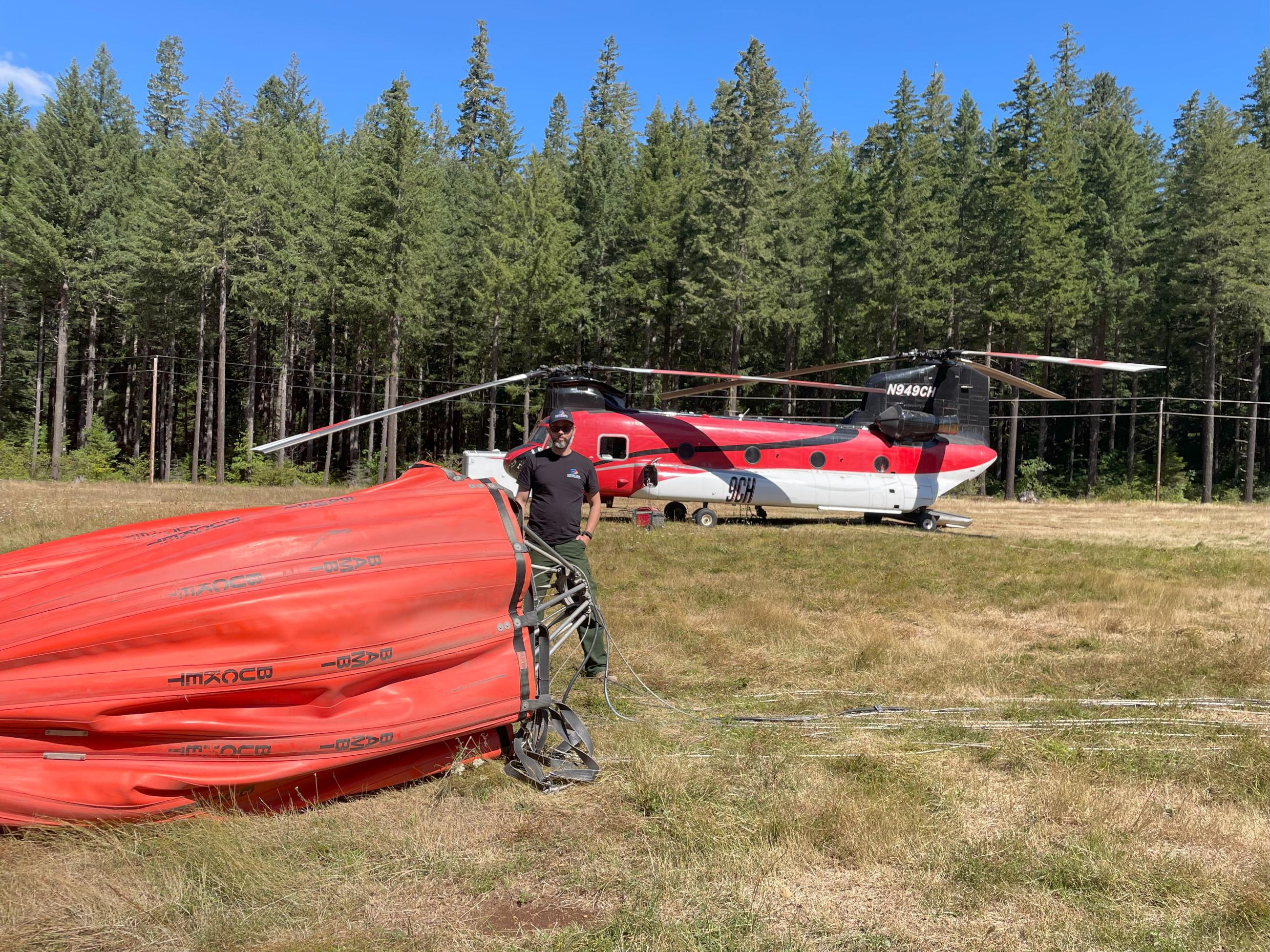 Air Ops Trainee Caleb Schreiber stands beside a 2,600 gallon Bambi Bucket carried under a Chinook Helicopter. It is bright orange, helicopter in the background.