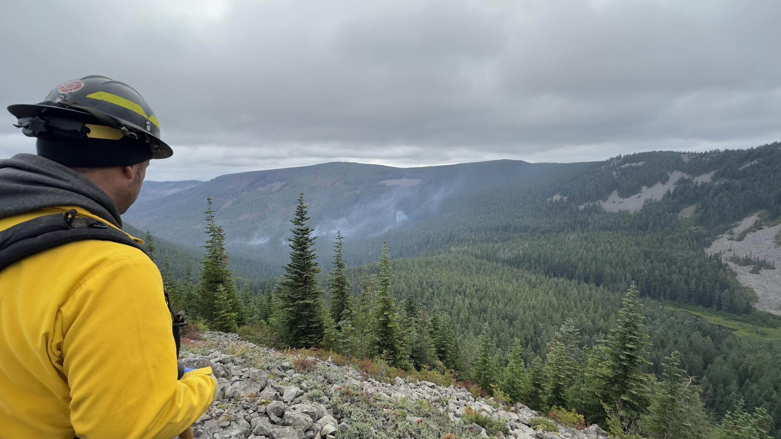 a firefighter is seen standing on a ridge line overlooking the fire area with smoke in the valley below