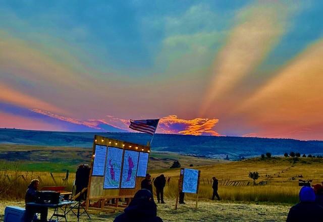 Photo of a sunrise behind an American flag which is attached to an information board out in a field of tall, brown grass. 